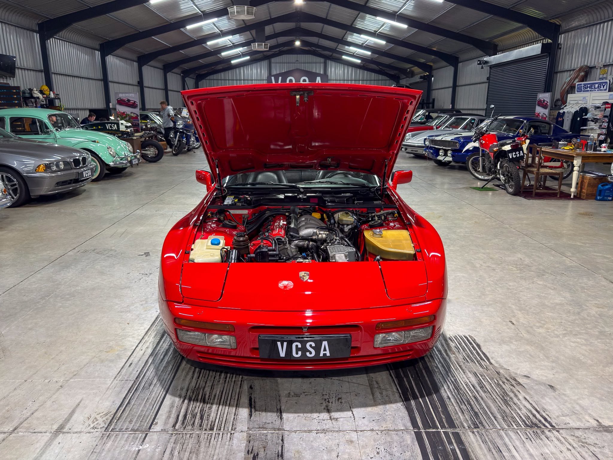 Red sports car with its hood raised in a tidy garage filled with vintage cars and motorcycles in the background, engine visible in foreground.