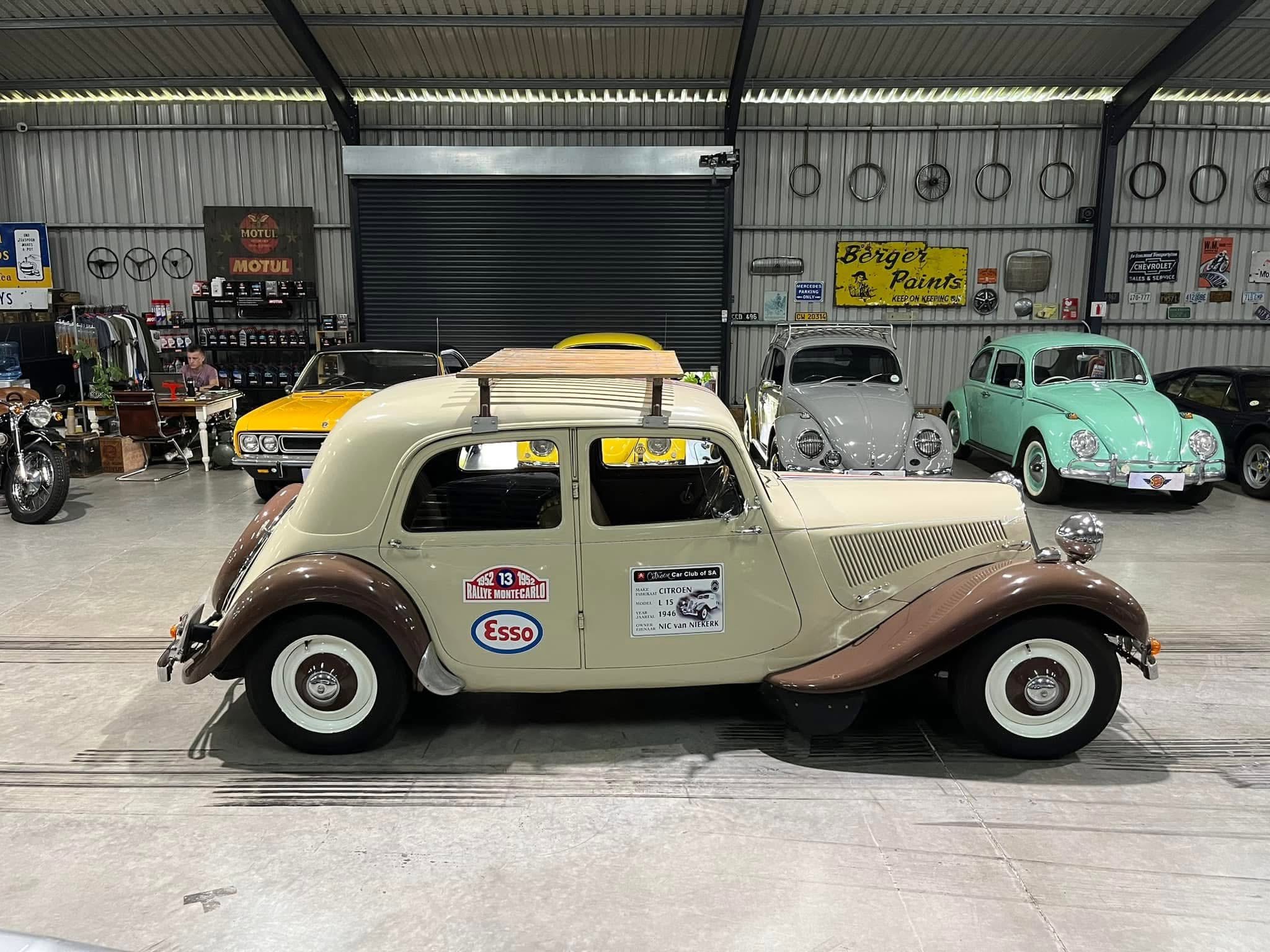 Inside a retro car showroom with vintage cars parked side by side, beige car in foreground and a yellow sports car behind it; walls lined with old signs and circular hubcaps.