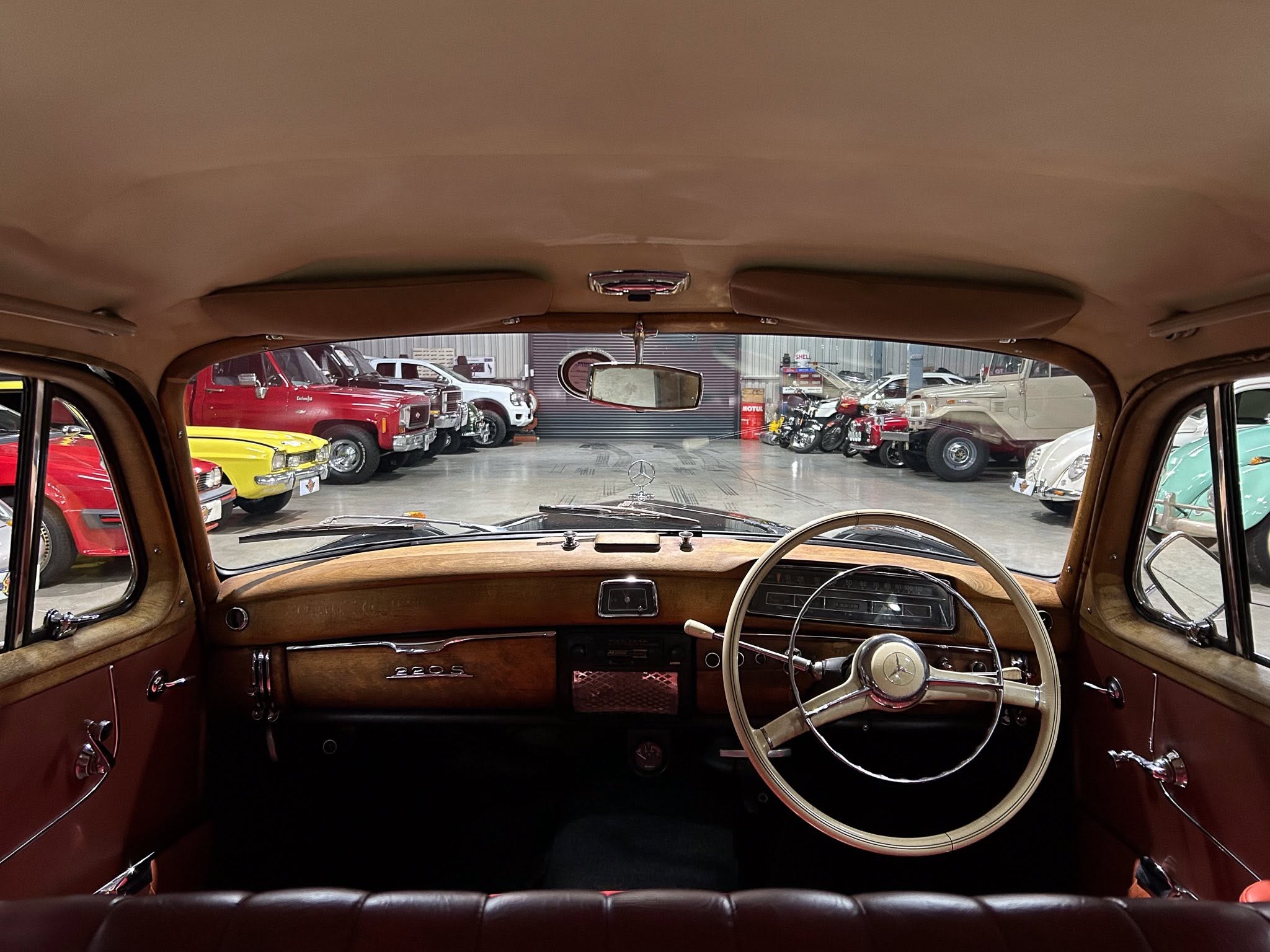 View from the back seat of a brown classic car with tan headliner; showroom of vintage cars visible through the windshield.Good for context: interior focus with outside showroom.