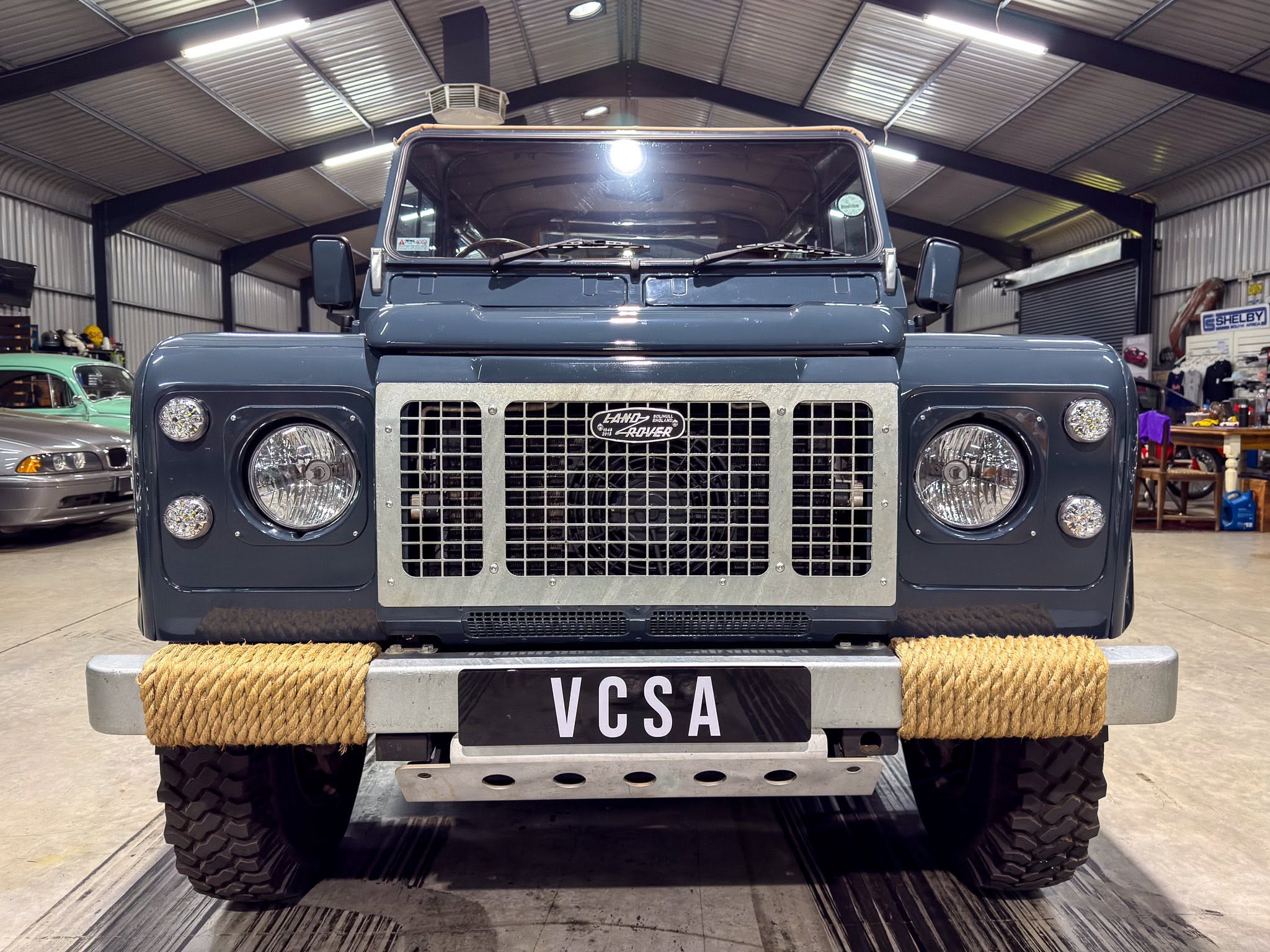 Front view of a vintage blue Land Rover inside a workshop, with a rope-wrapped bumper and large metal grille.](https://)