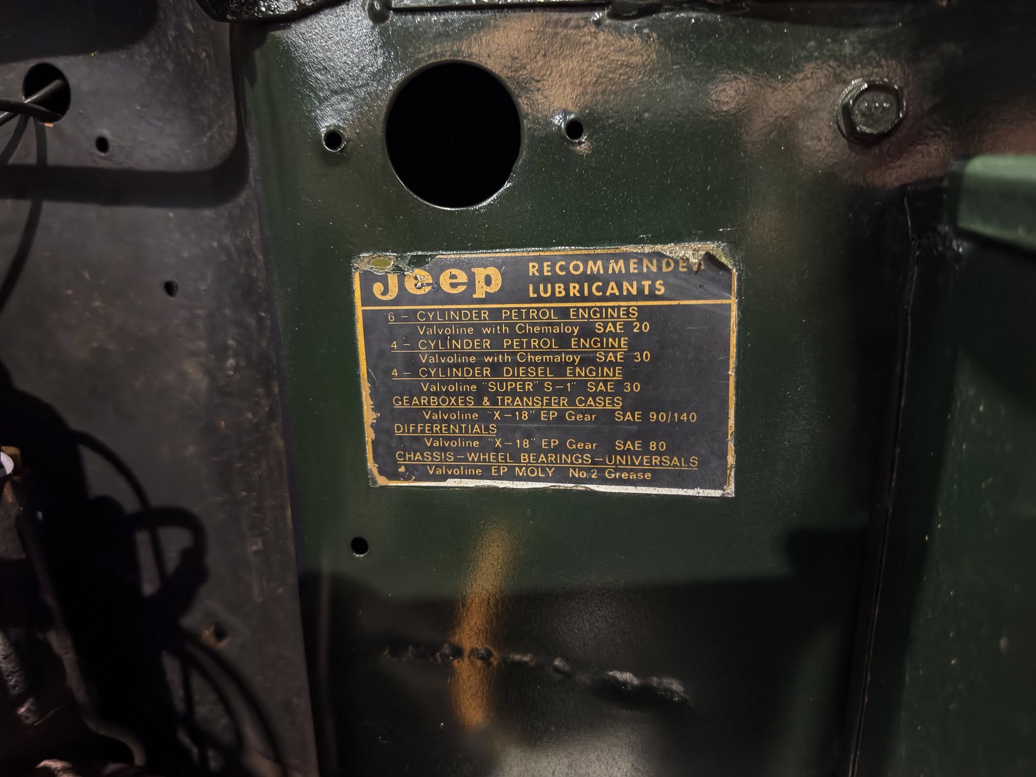 Close-up of a green vehicle engine bay with a yellow-and-black plate reading 'Jeep Recommended Lubricants' and a circular hole above it.