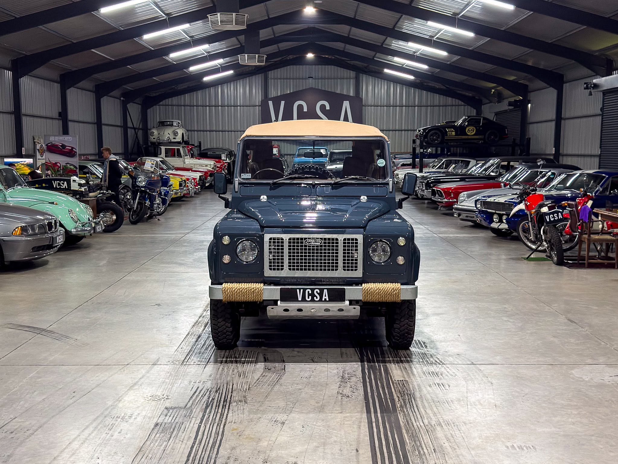 Front view of a blue vintage SUV with beige top inside a large indoor classic car showroom, flanked by rows of colorful vintage cars.