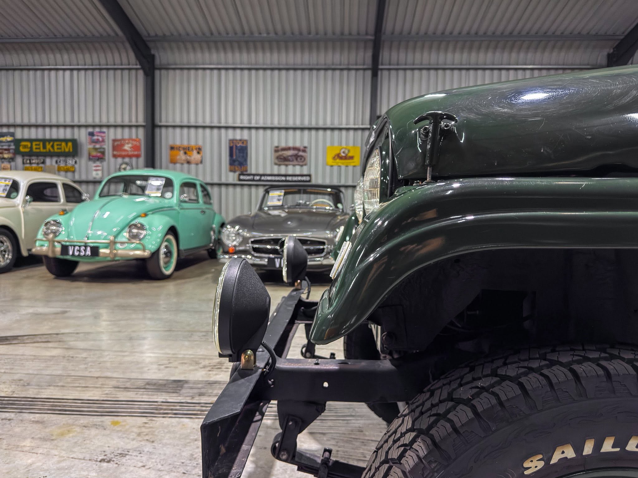 Close-up of a dark green vintage car’s front fender and wheel, with other classic cars and retro signs in a showroom background.