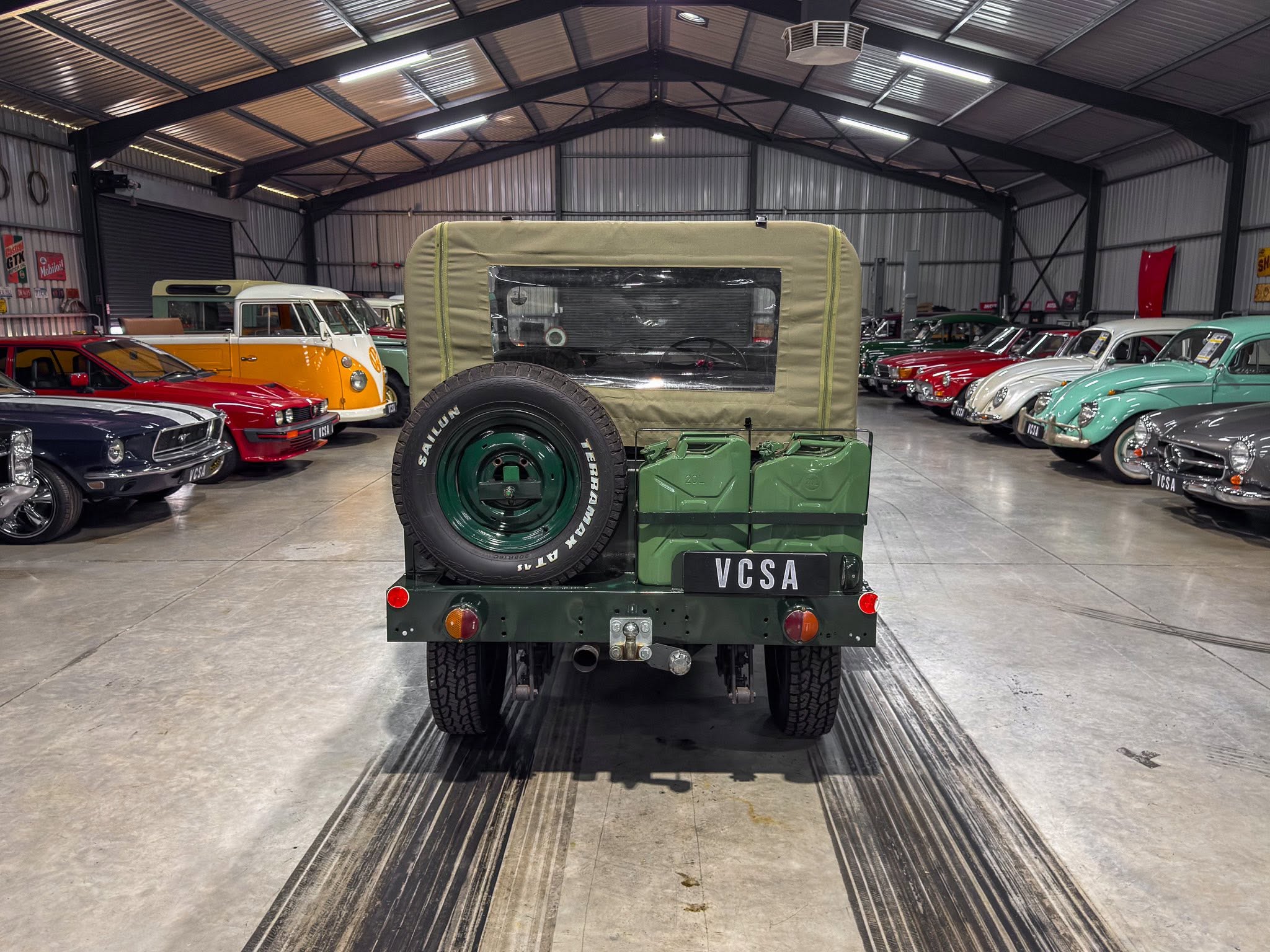 Rear view of a vintage military jeep in a showroom, with classic cars lining both sides in a metal-walled warehouse.
