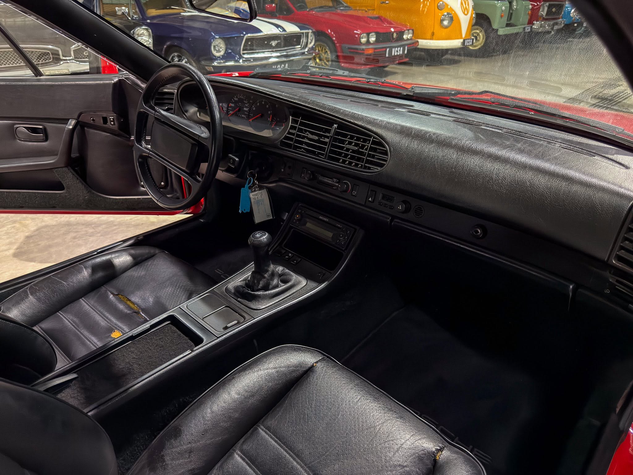 Inside a classic car: black leather seats, manual gear shifter, and a dashboard with analog gauges; row of vintage cars visible through windshield.