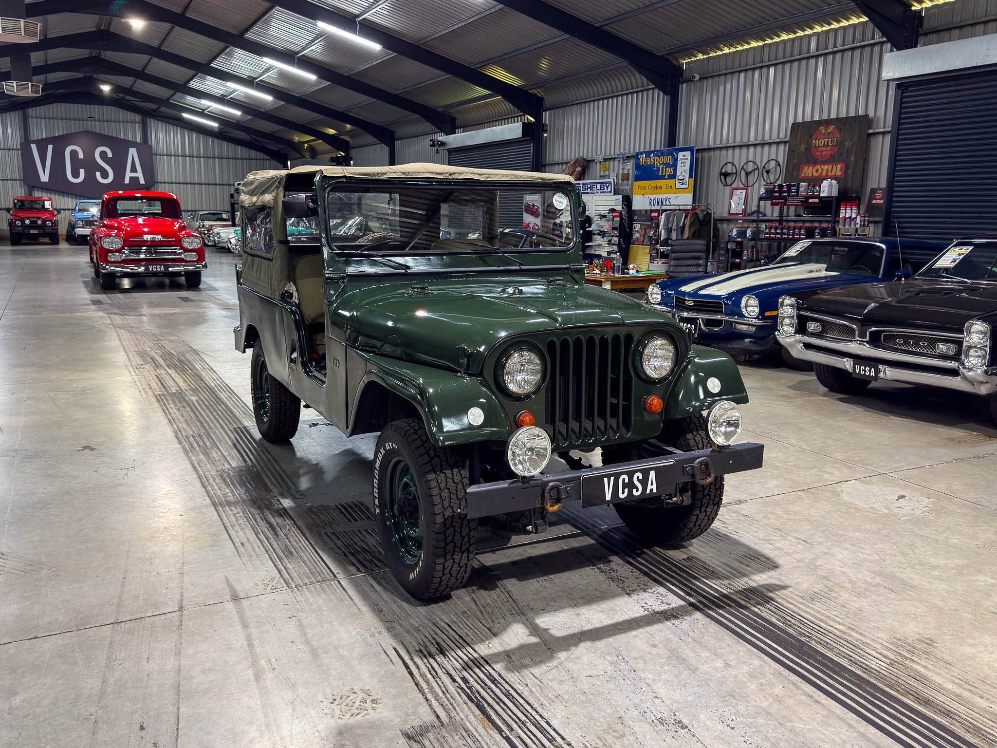 Indoor showroom with vintage cars, green military Jeep in foreground and red classic car in background; large VCSA sign on wall.