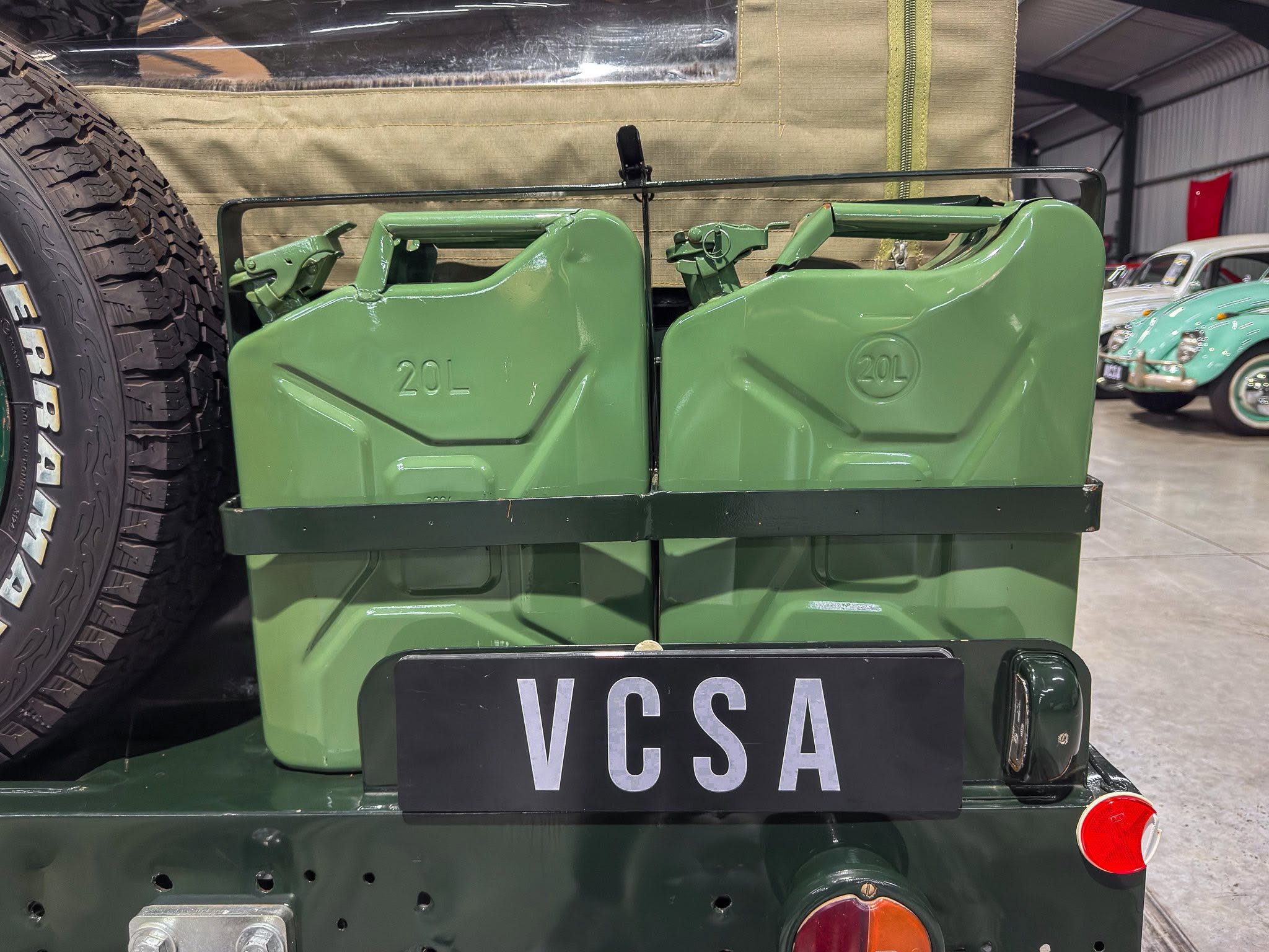Rear view of a dark-green military-style vehicle with two large green jerry cans mounted on the back and a spare tire on the left; license plate reads VCSA.