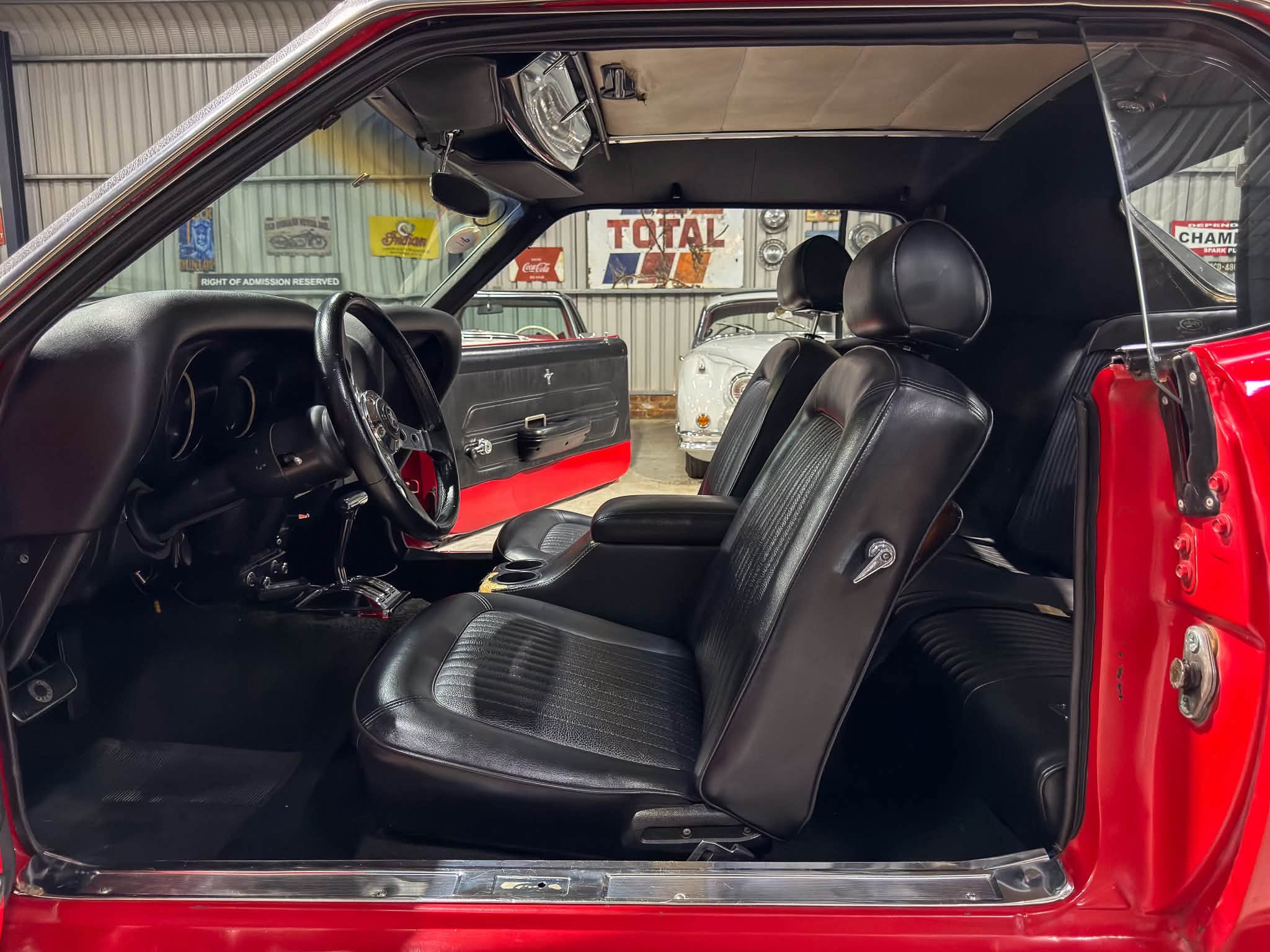 Open-door view of a red vintage car interior with black leather seats and exposed headliner in a garage backdrop with vintage signs.