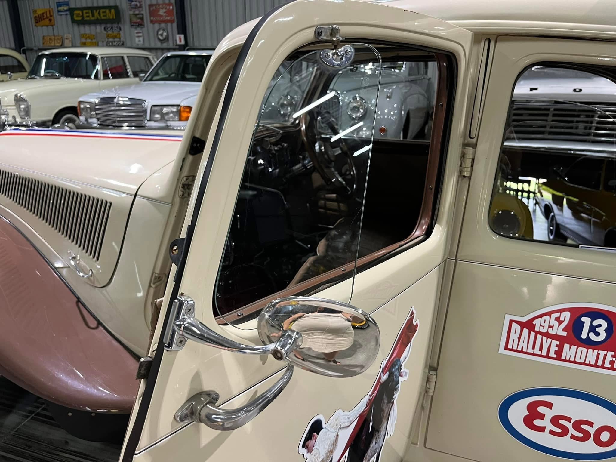 Close-up of a beige vintage car door with a chrome side mirror, showing the interior dashboard; a Rallye Monte Carlo sticker is on the door with other classic cars in the background in a showroom.