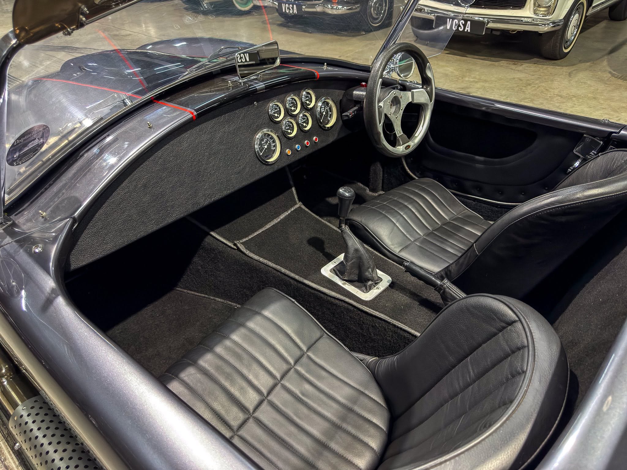 Cockpit of a vintage sports car with a cluster of circular gauges, black leather seats, and a silver steering wheel.