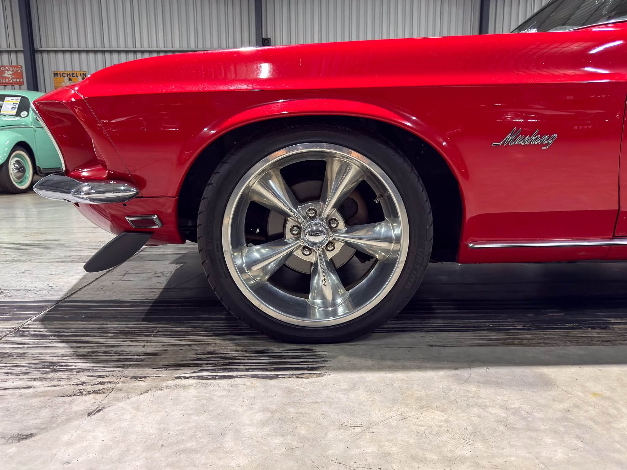 Close-up of a red Mustang fender and wheel in a showroom, highlighting chrome wheel and body styling.
