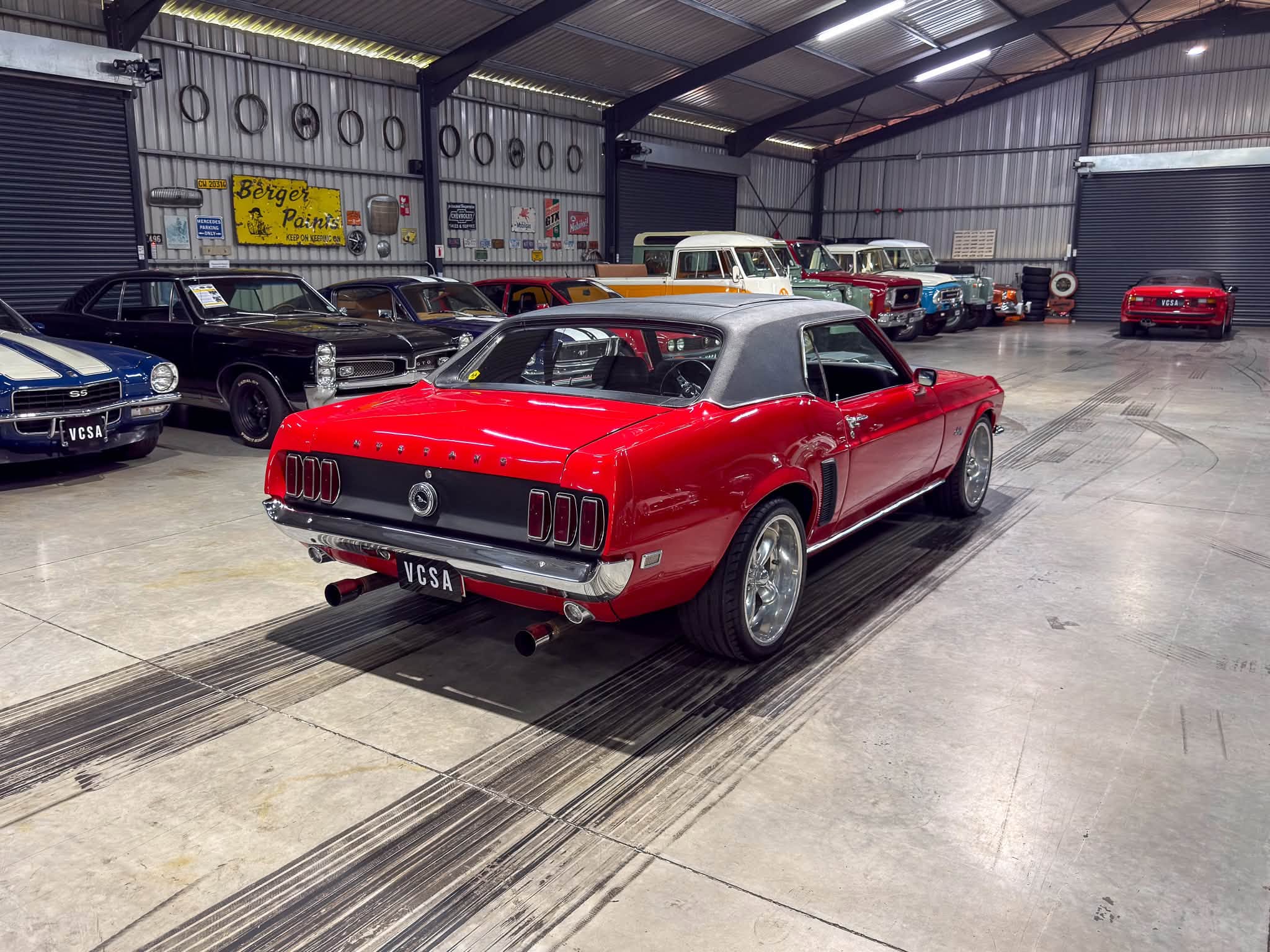 Inside a classic car showroom with red convertible in foreground and vintage cars lined along metal walls.