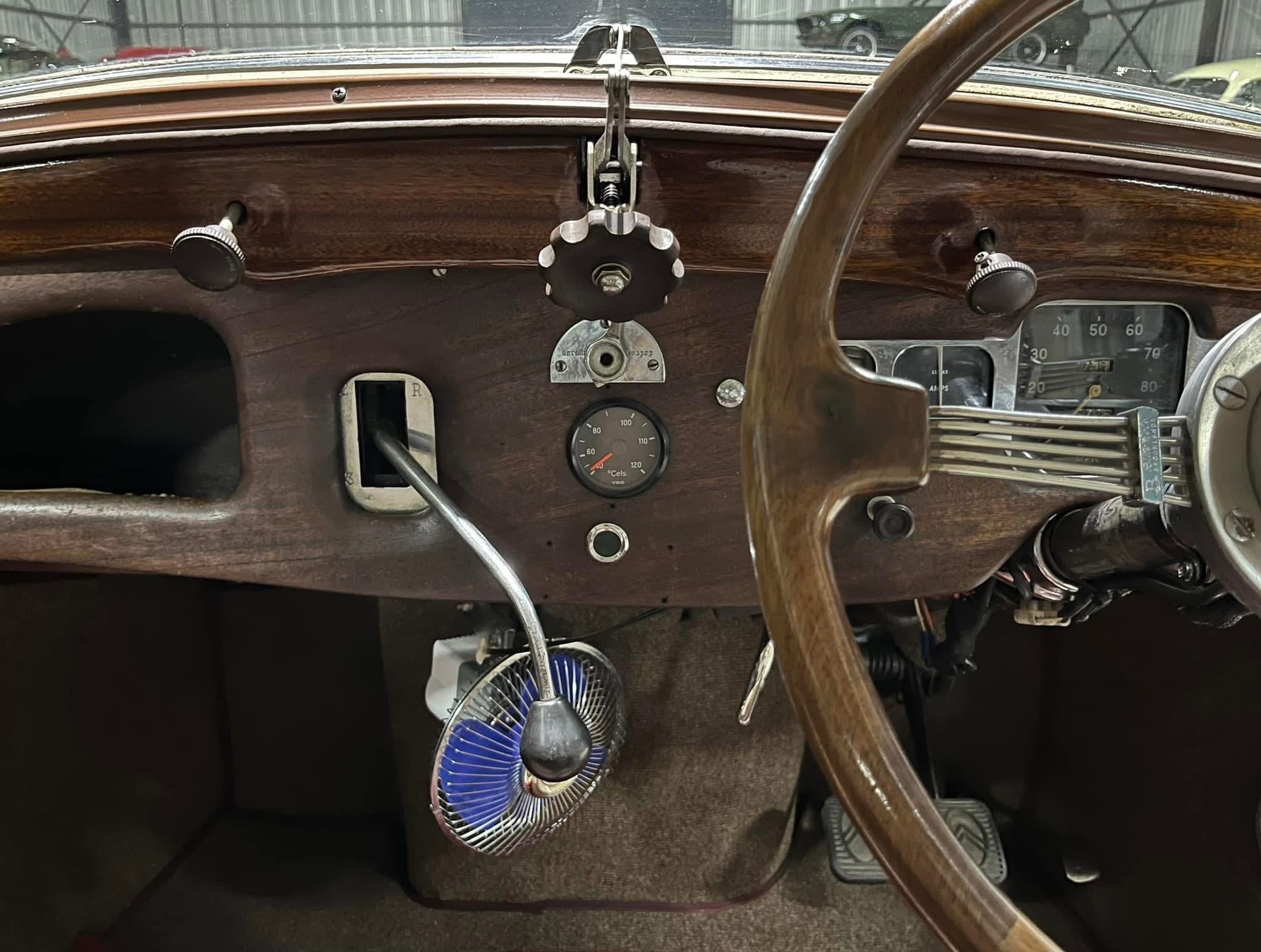 Interior of a vintage car cockpit with a wooden dash, large steering wheel, and analog gauges; a blue portable fan sits on the floor behind the pedals.
