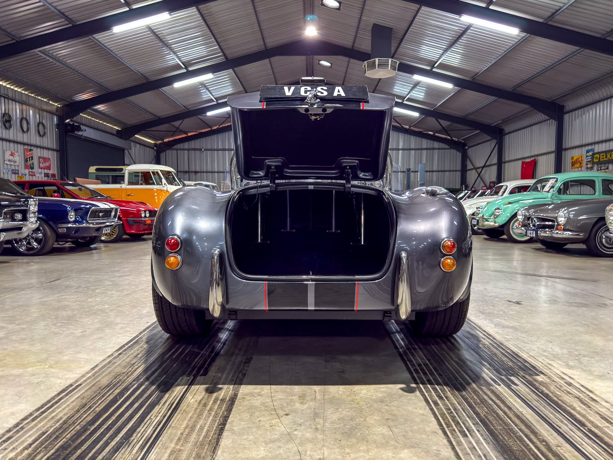 Rear view of a glossy vintage car with its trunk open in a classic car showroom, surrounded by other colorful older automobiles.
