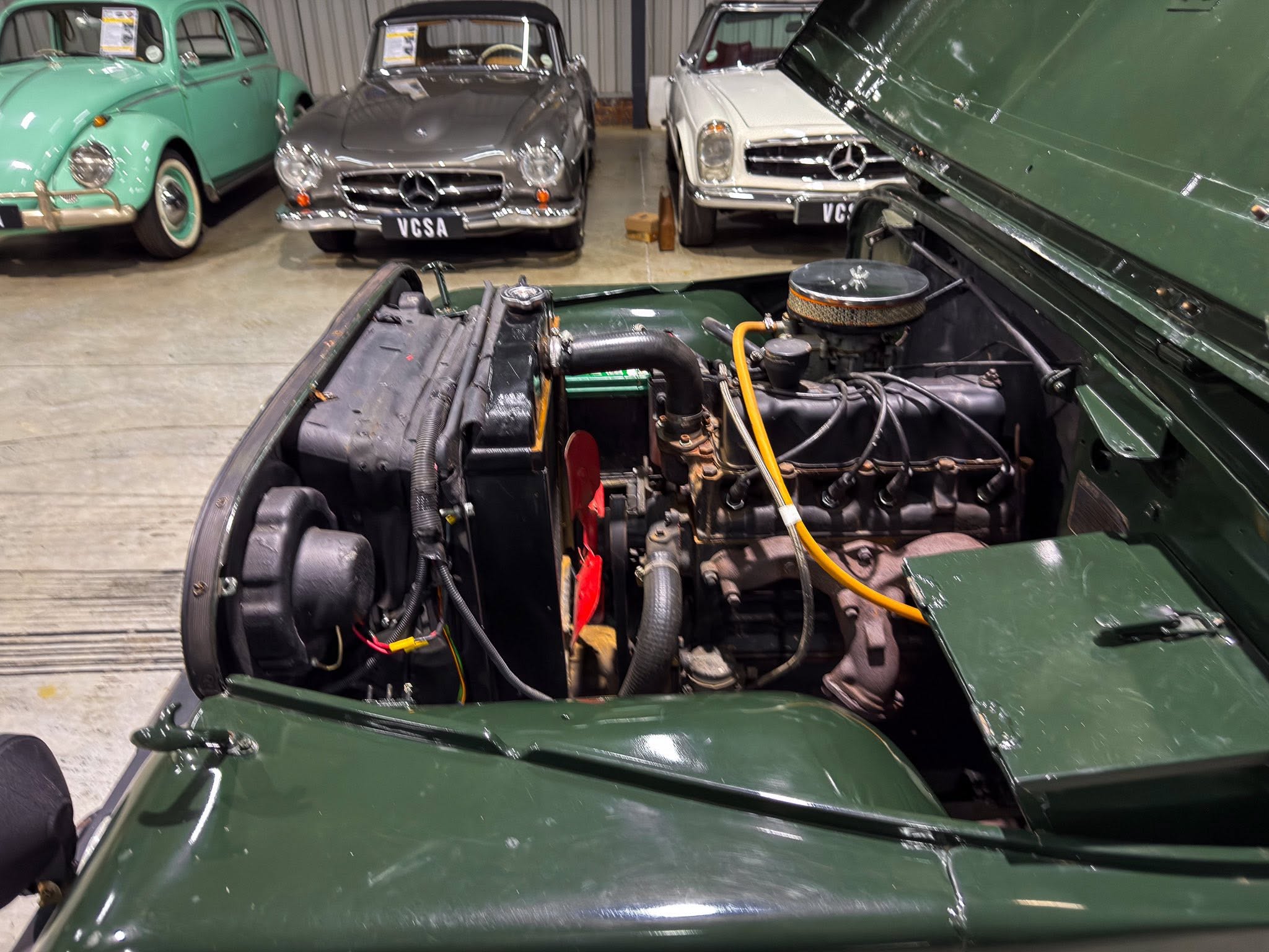Open engine bay of a dark green classic car, with vintage cars in a storage garage behind it.