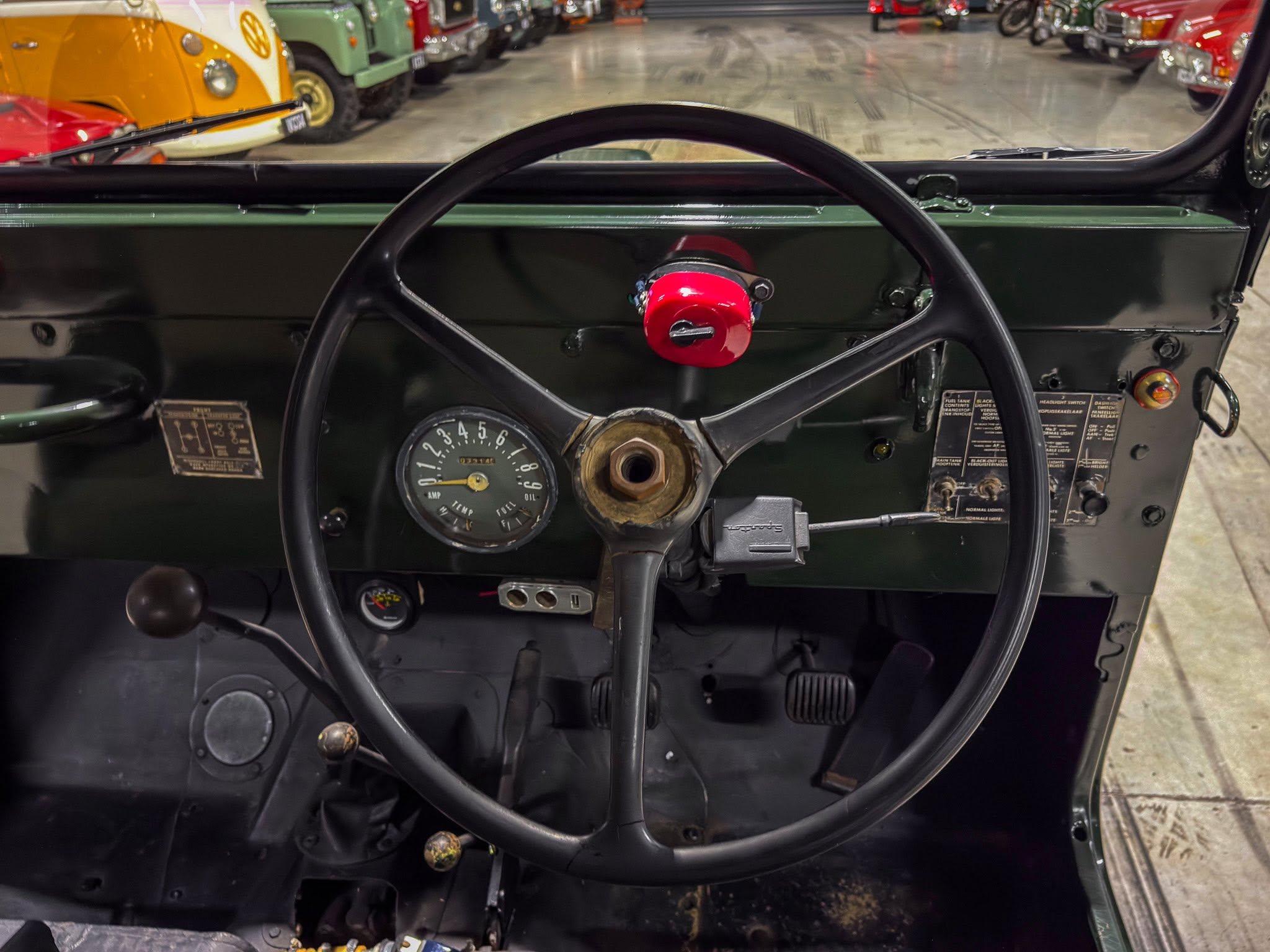 Inside a vintage car cockpit, showing a large steering wheel, red horn cap, gauge cluster, and old controls with a row of classic cars visible through the windshield.