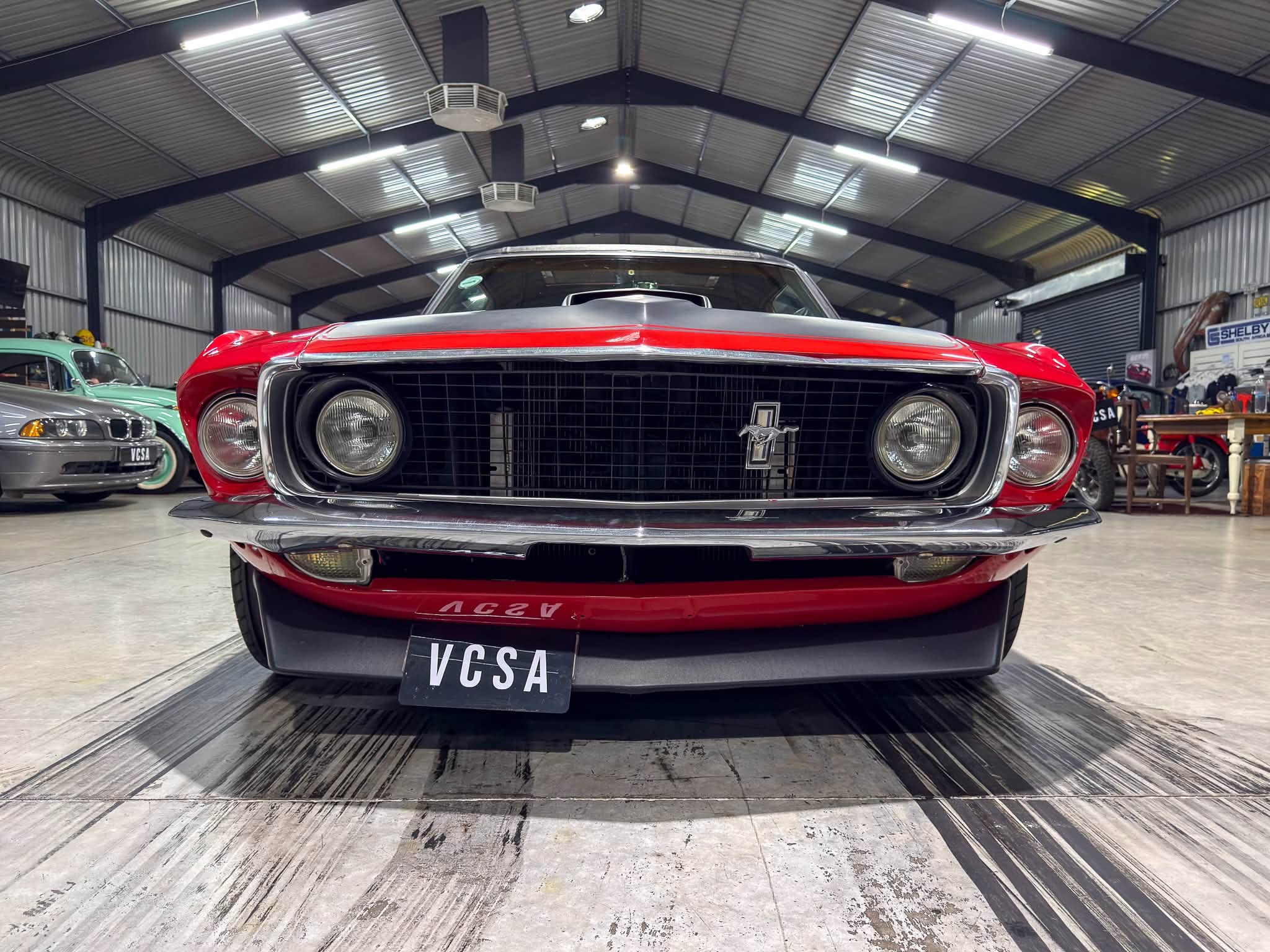 Low-angle front view of a red vintage muscle car with chrome grille and round headlights in an indoor showroom, license plate VCSA.