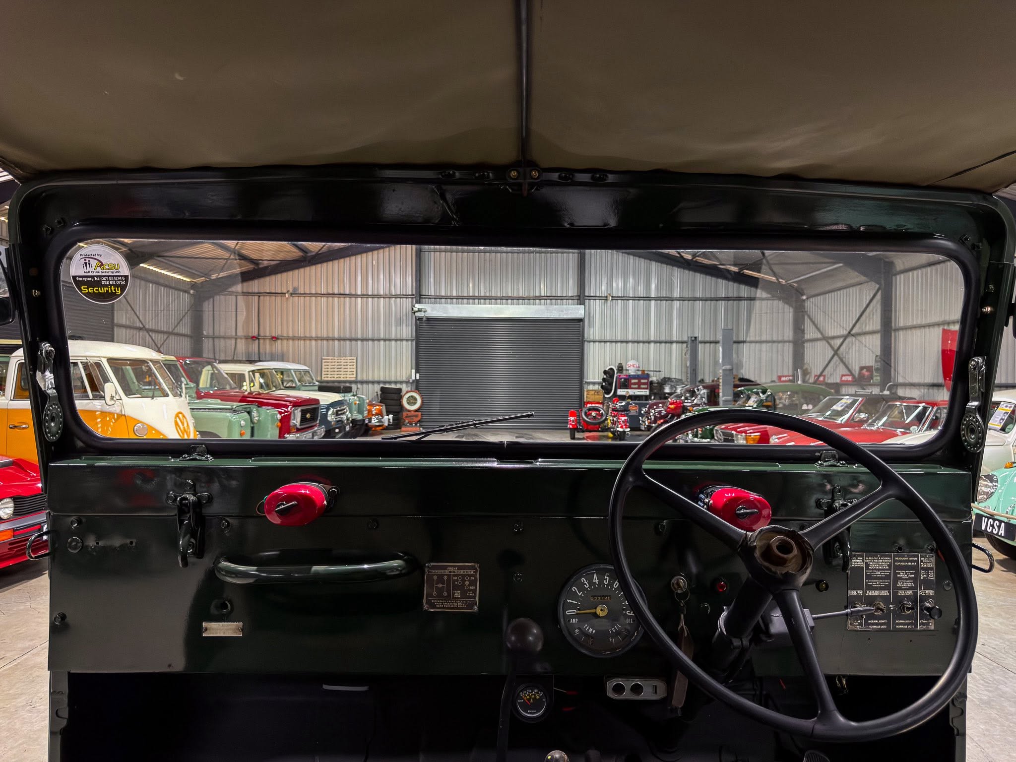 View from inside a vintage car showing the dashboard, steering wheel, and a showroom filled with classic cars beyond the windshield.