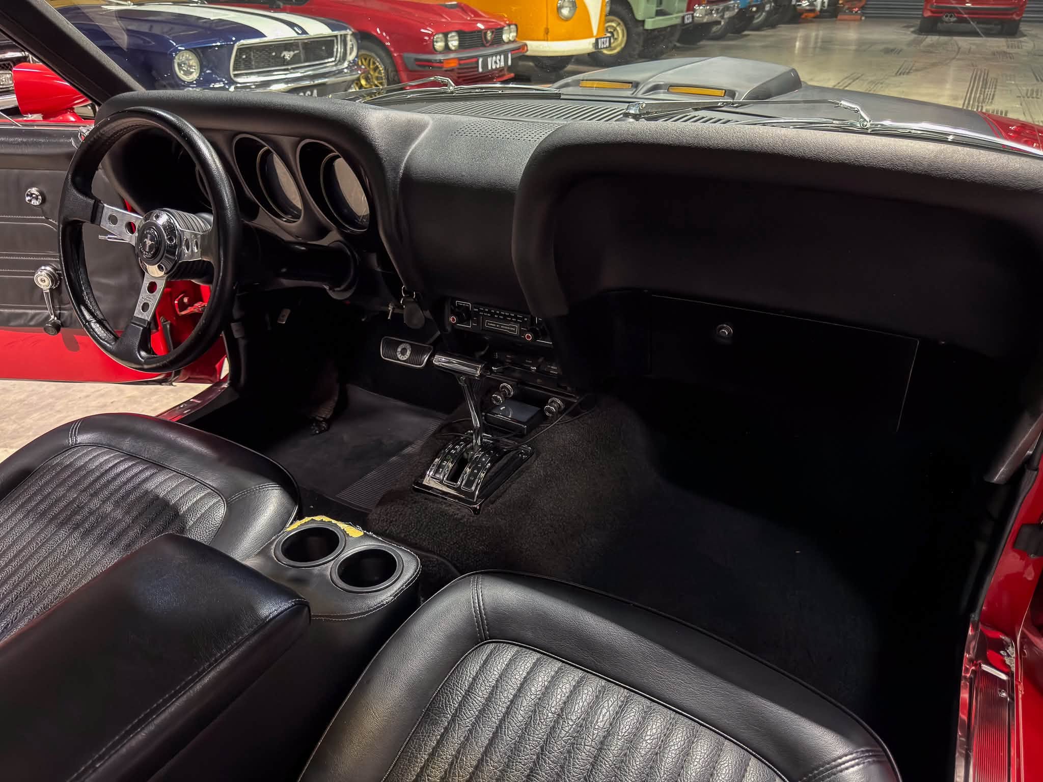 Interior of a vintage car with black leather seats, three-spoke steering wheel, and gauges, showroom of classic cars visible through the windshield