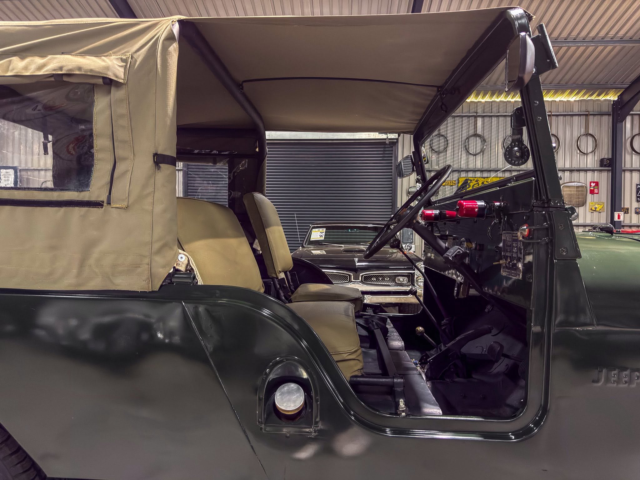 Interior view of a dark green vintage jeep with a tan canvas top raised, parked inside a metal workshop or garage.