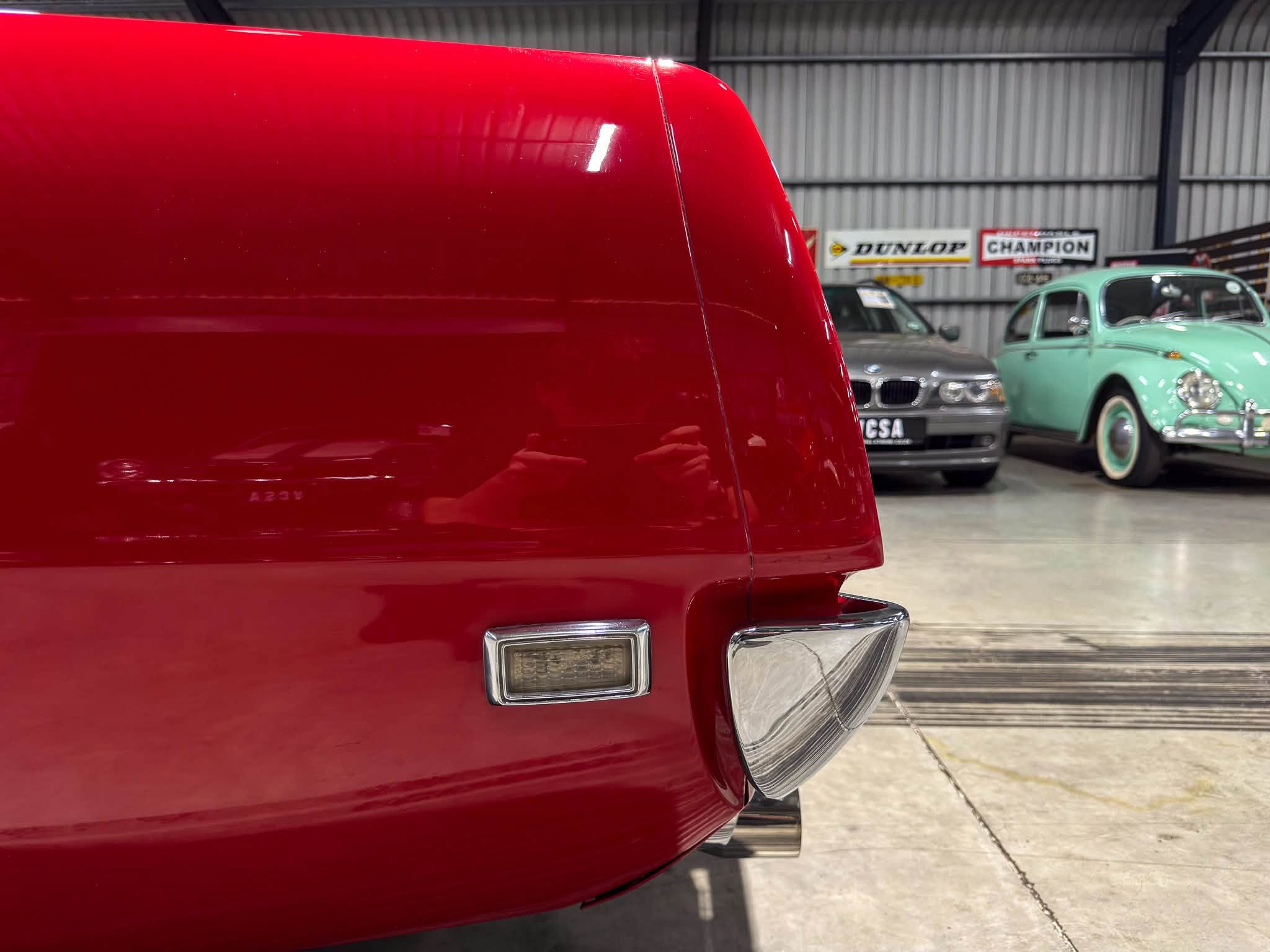 Close-up of the rear corner of a glossy red classic car, with chrome bumper and taillight, in a showroom with other vintage cars in the background.
