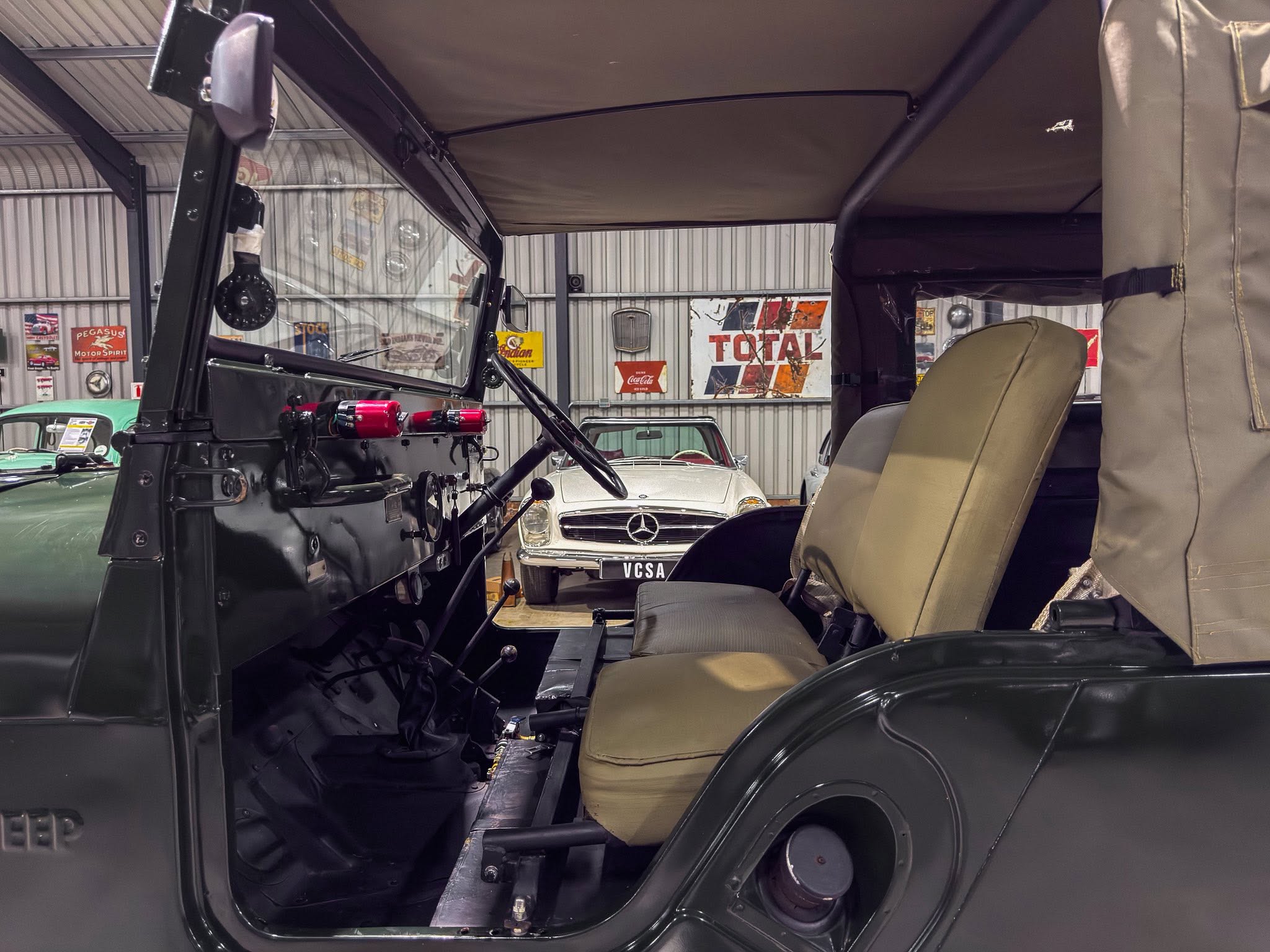 Interior view of a dark green vintage jeep with the canvas top open, in a museum showroom with classic cars and retro posters in the background.