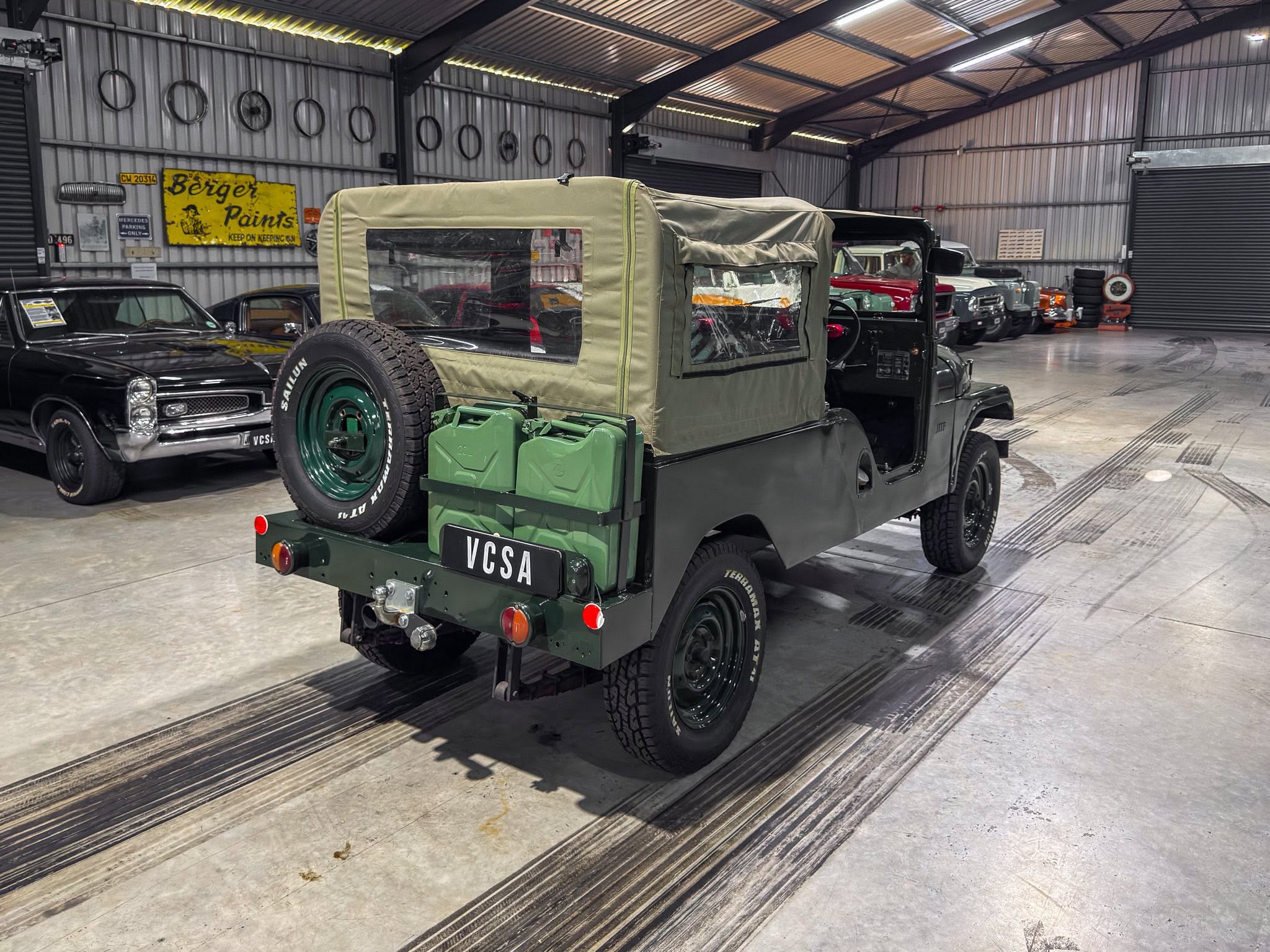 Rear view of a green vintage Jeep with a tan canvas top in a showroom, surrounded by other classic cars and signs on the wall.