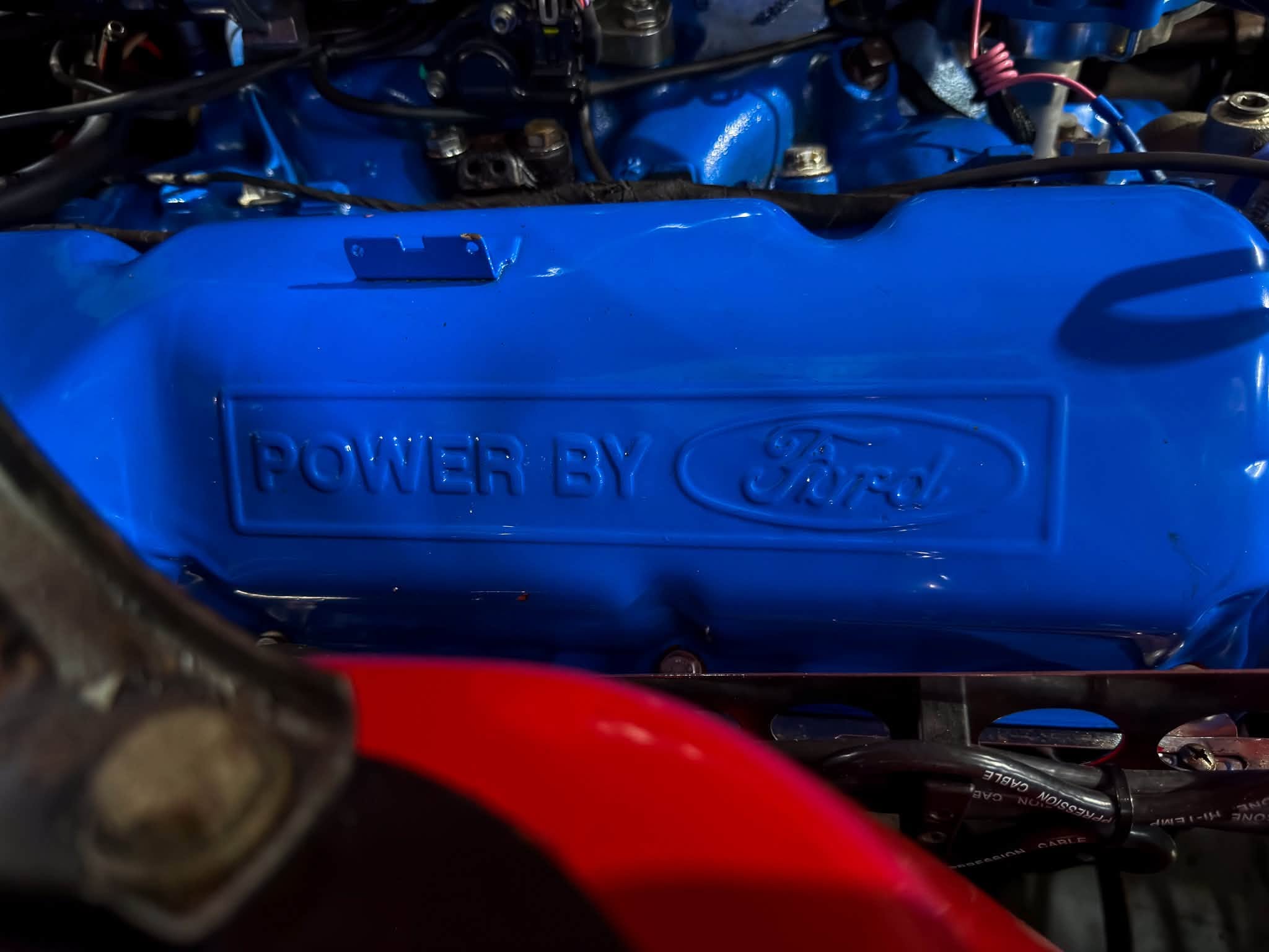 Blue engine bay close-up showing a blue valve cover with embossed 'POWER BY Ford' and a red body panel in the foreground.