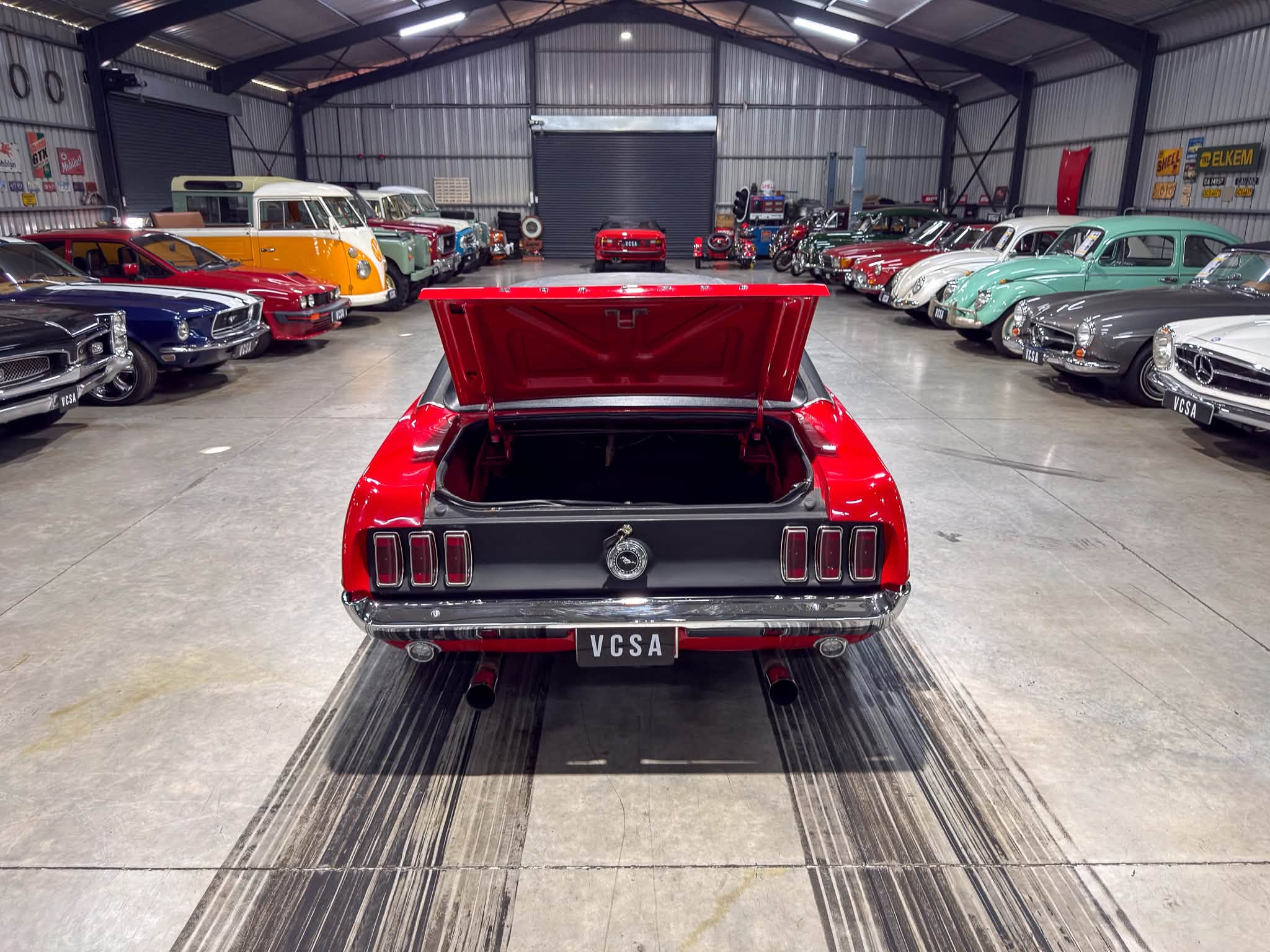 Rear view of a red classic car with its trunk open, in a spacious showroom filled with vintage cars on both sides.