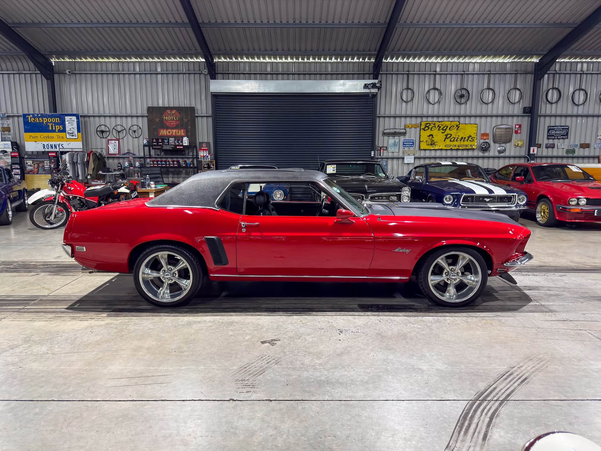 Red vintage convertible with a gray hardtop parked inside a showroom-like garage, surrounded by other classic cars and retro signs on corrugated metal walls.