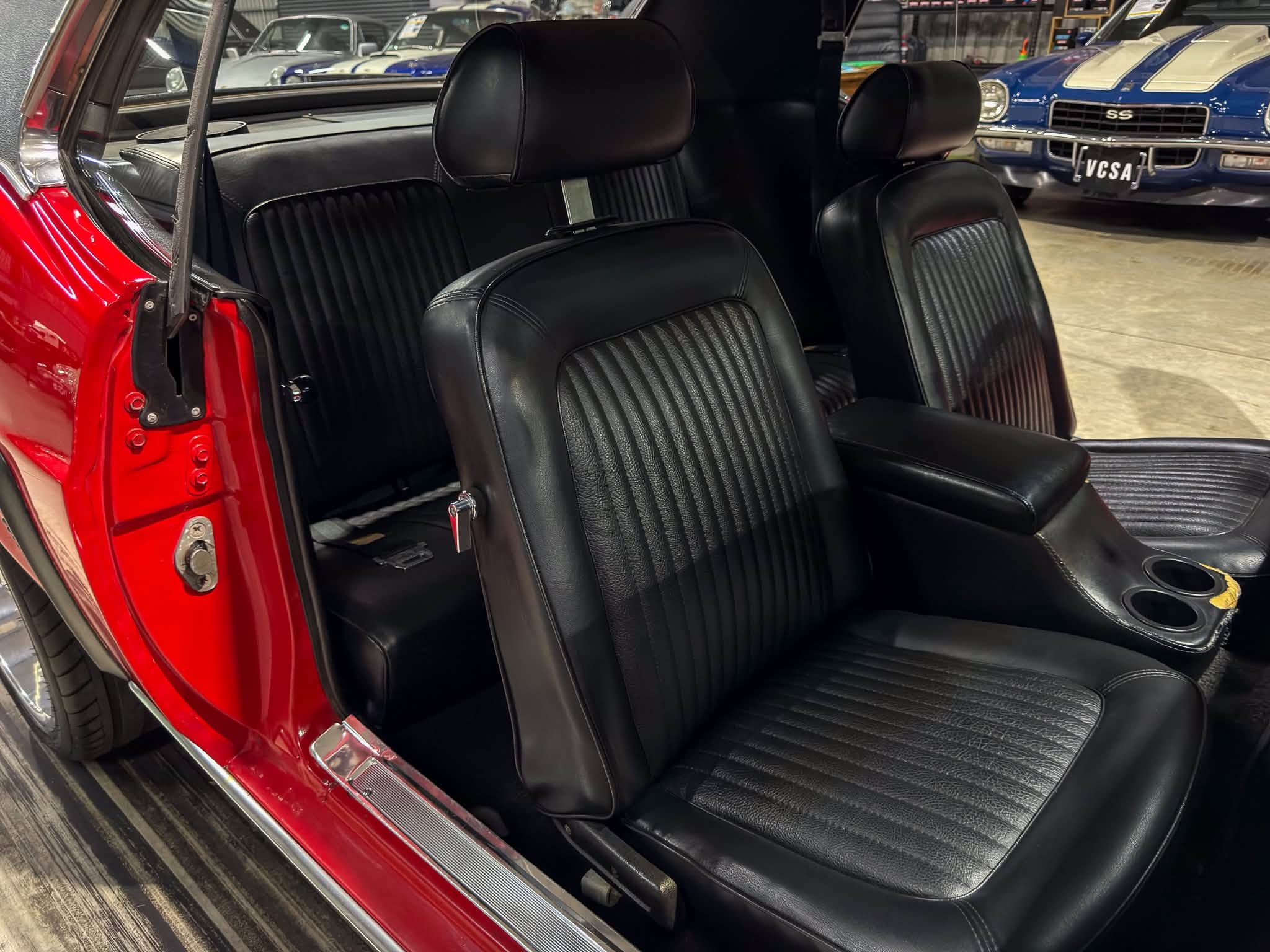 Red car interior with black leather seats, showing front bucket seat and rear bench in a showroom. Outside, other classic cars are visible.