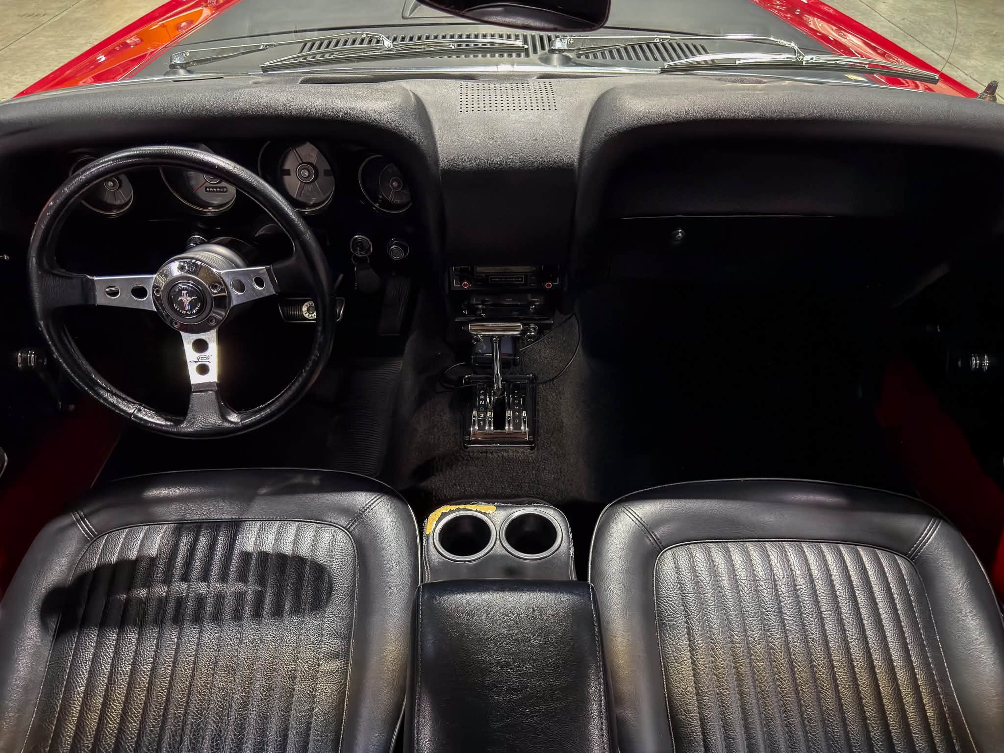 Front-seat interior of a classic car: black leather seats, dashboard with analog gauges, and a chrome steering wheel seen from the back seats.