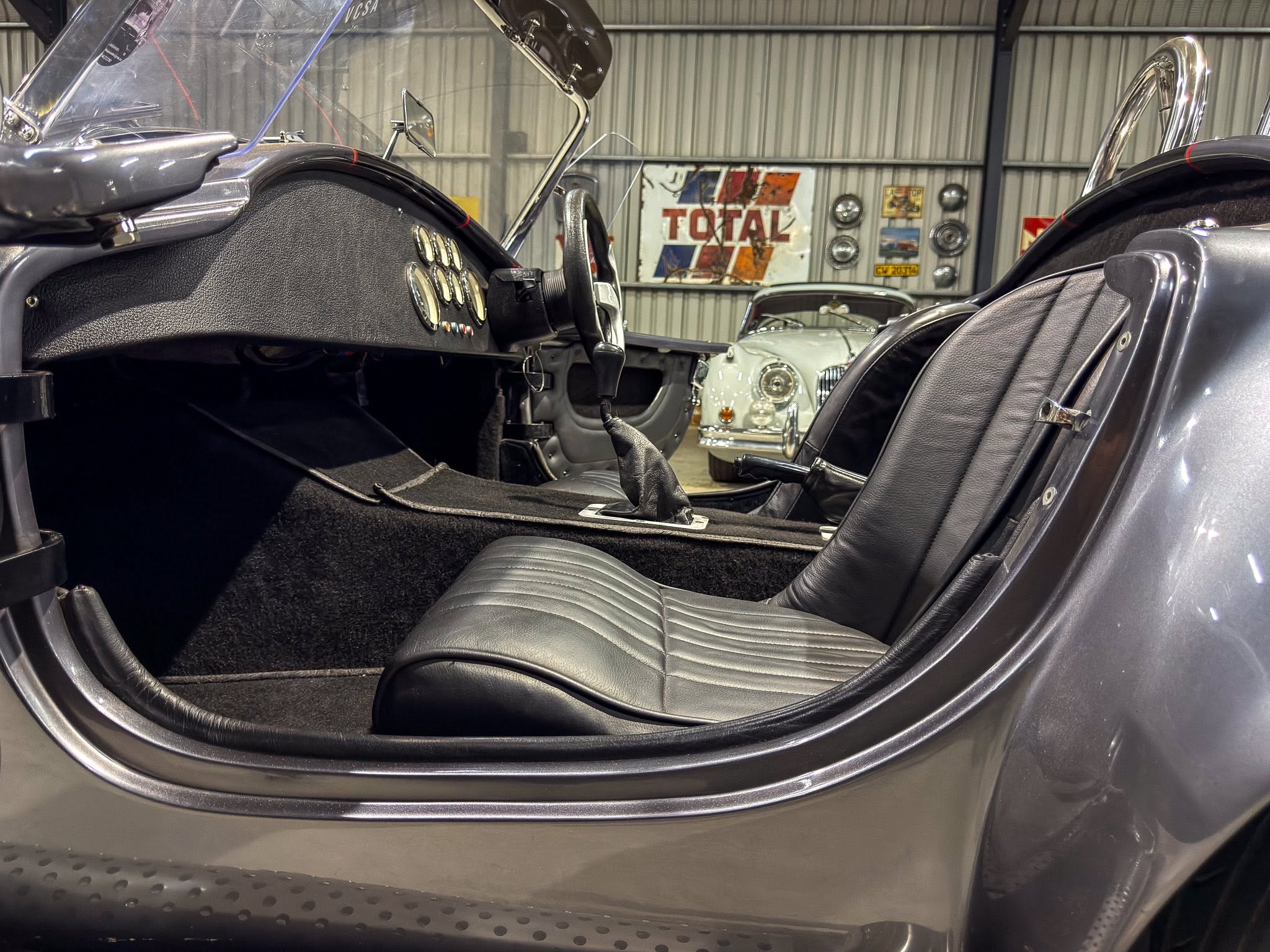 Interior view of a vintage car: black dashboard with analog gauges and a slim steering wheel, leather seats, in a showroom setting with another classic car in the background