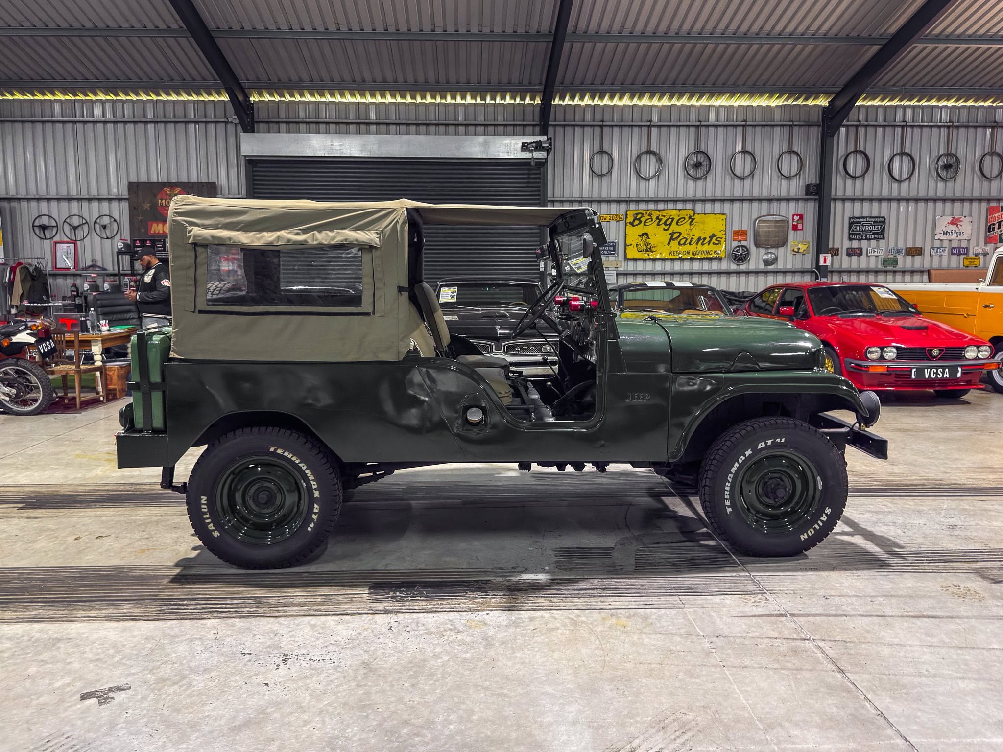Green military jeep with a canvas top parked inside a spacious workshop, alongside vintage cars and retro signs on metal walls.