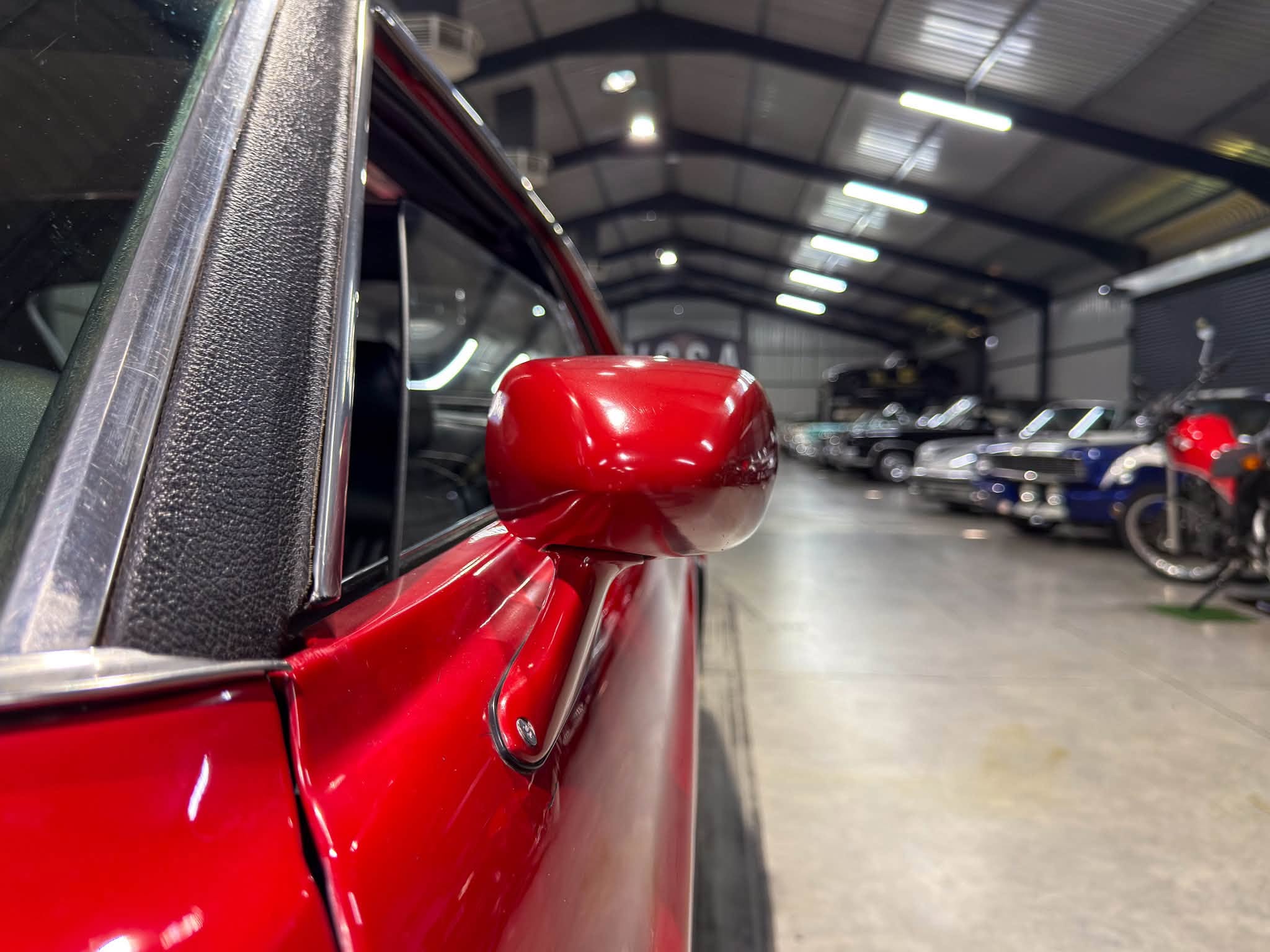 Close-up of a red car’s side mirror and door trim inside a spacious garage with other vehicles in the background