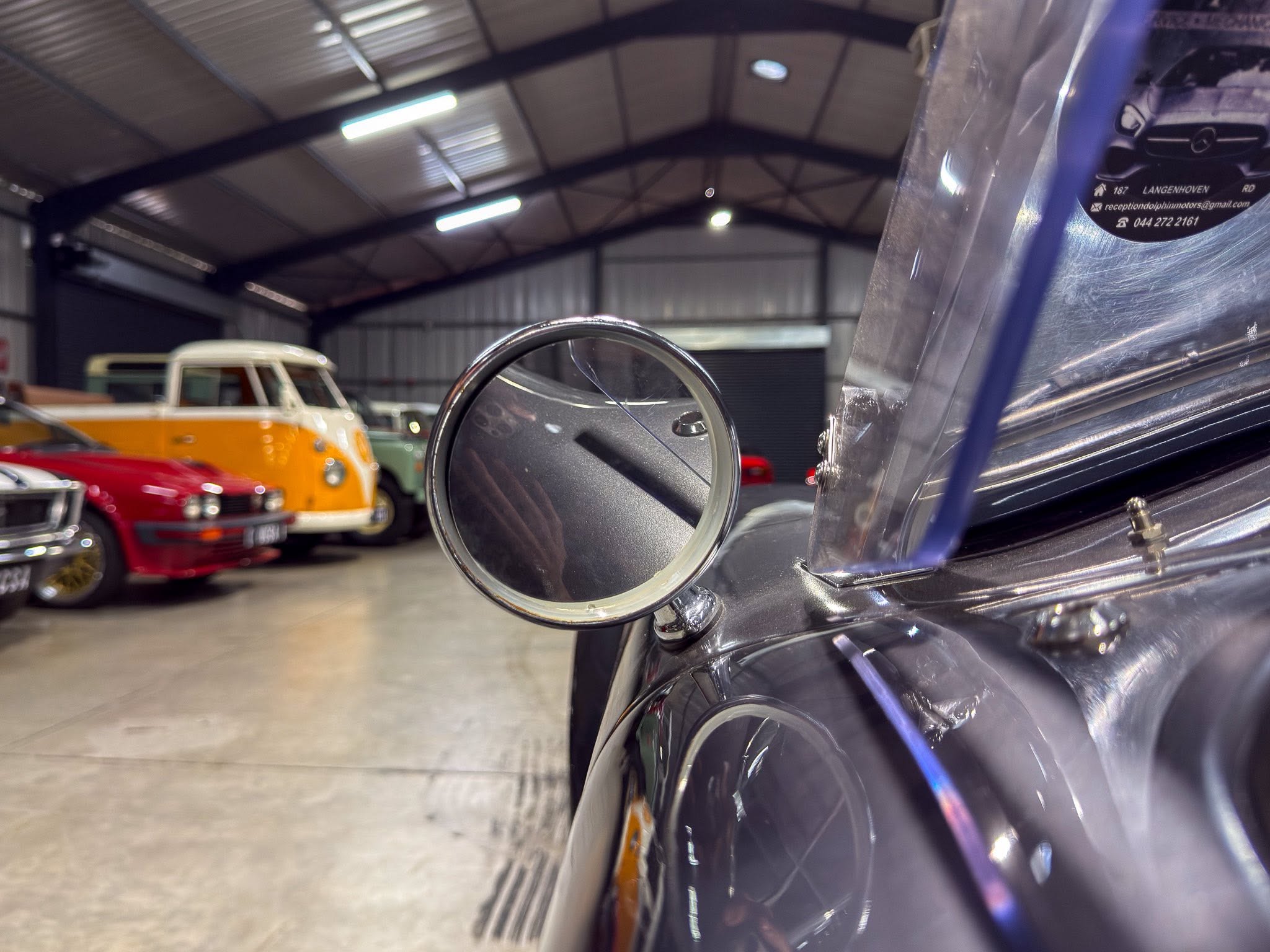 Close-up of a classic car's chrome headlight housing and fender in a workshop, with vintage cars blurred in the background.