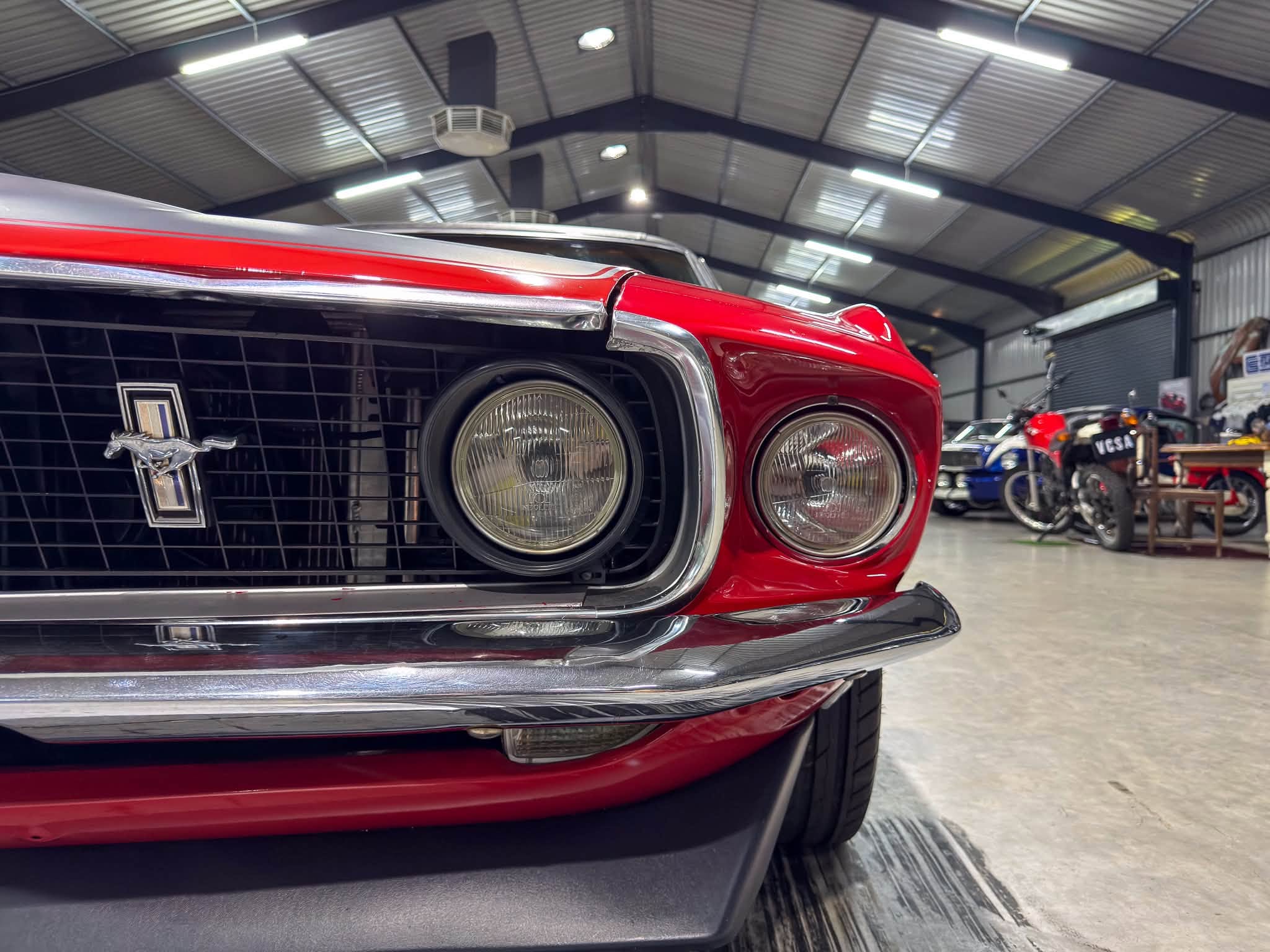 Close-up of the front end of a red vintage Ford Mustang with dual headlights and chrome grille in a workshop/showroom setting.