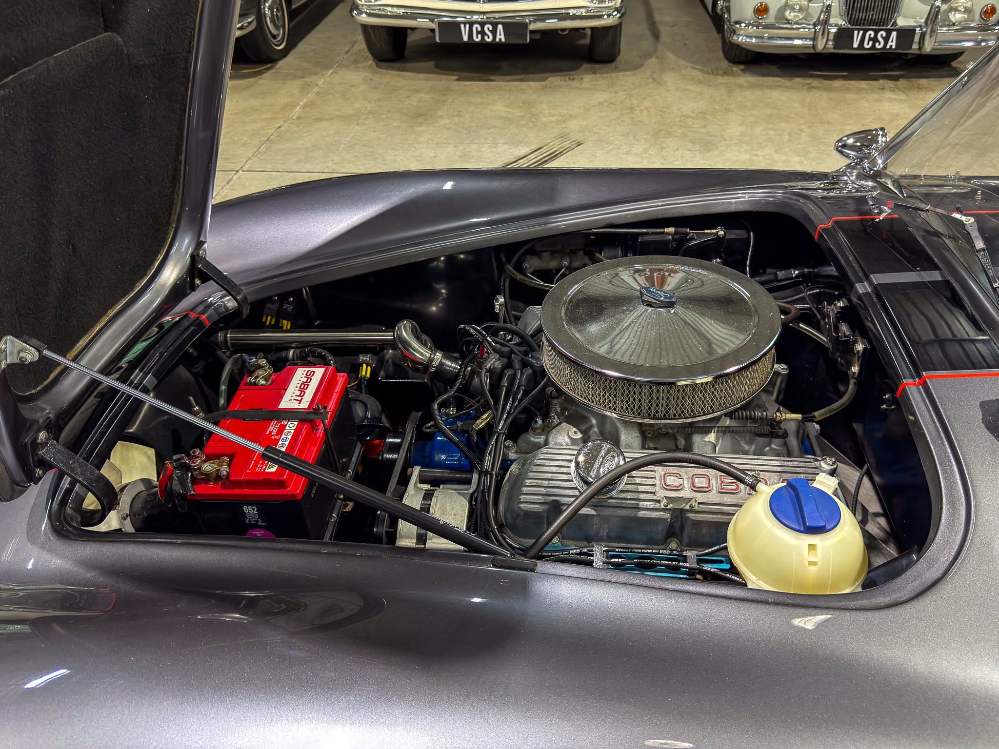 Open-hood view of a classic car engine bay with a large circular air filter, red battery, and surrounding components in a showroom floor.