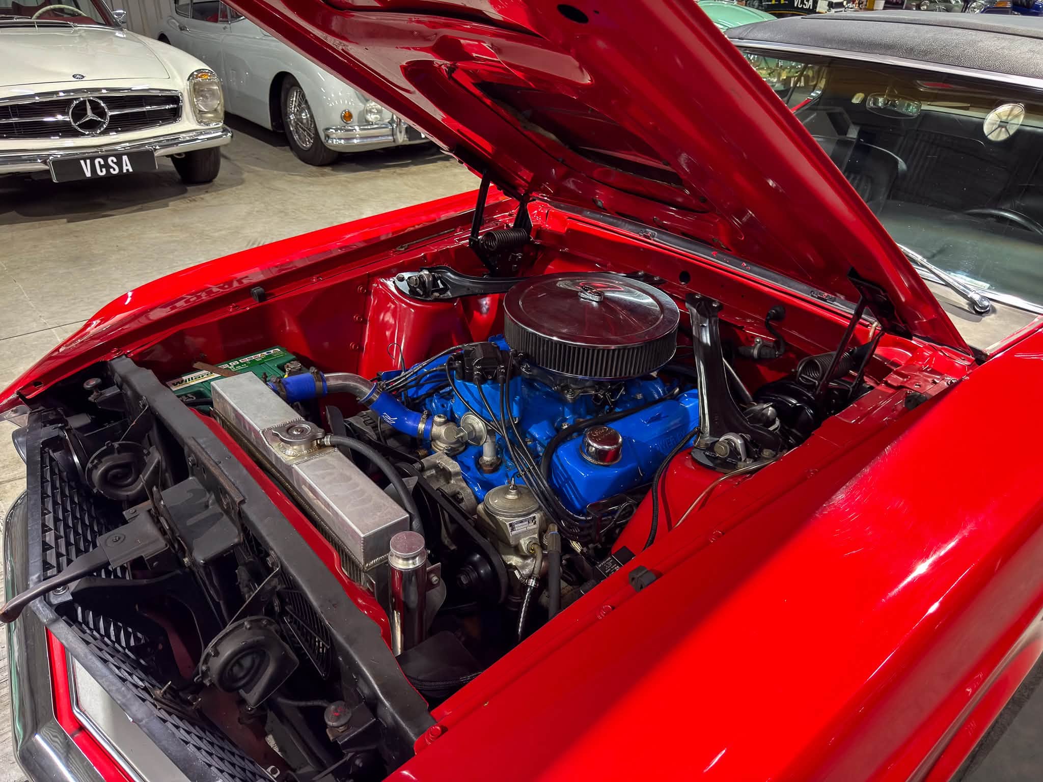 Red classic car with its hood raised, exposing a blue engine bay in a showroom with vintage Mercedes cars in the background.