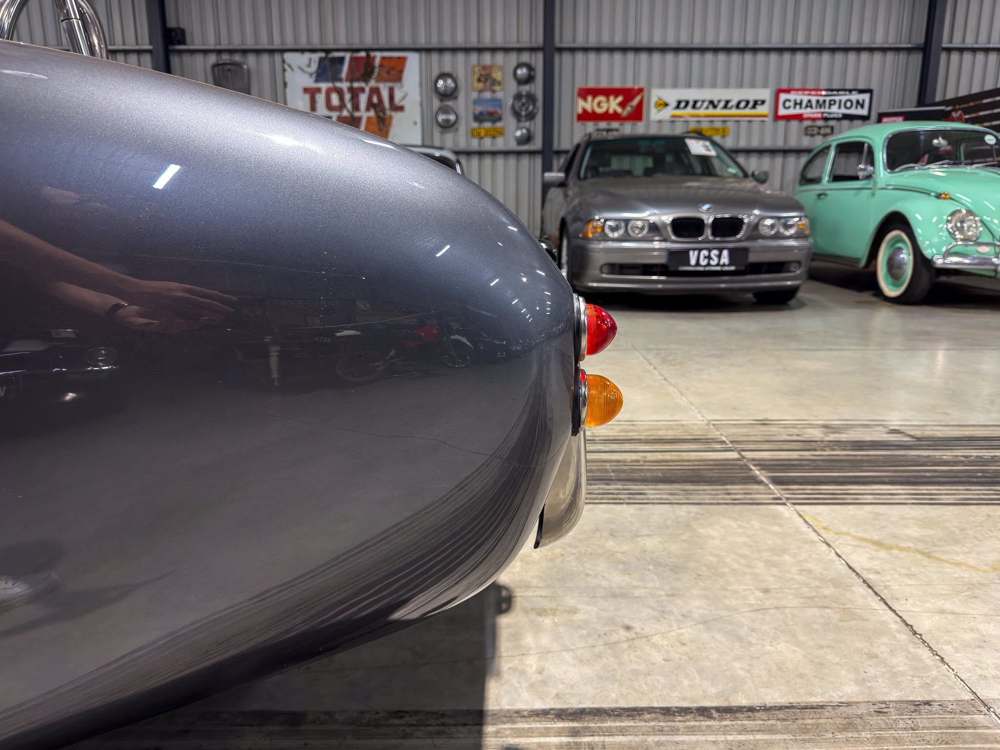 Close-up of a dark grey car's curved rear with a red tail light in a showroom or garage, other classic cars visible in the background.