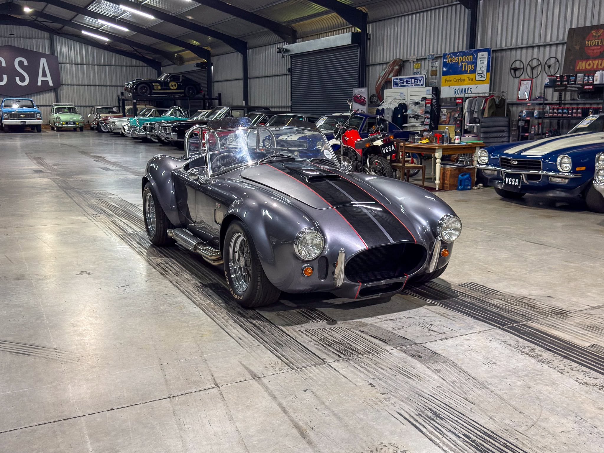 Grey vintage convertible sports car in a showroom with rows of classic cars and memorabilia in a warehouse setting.