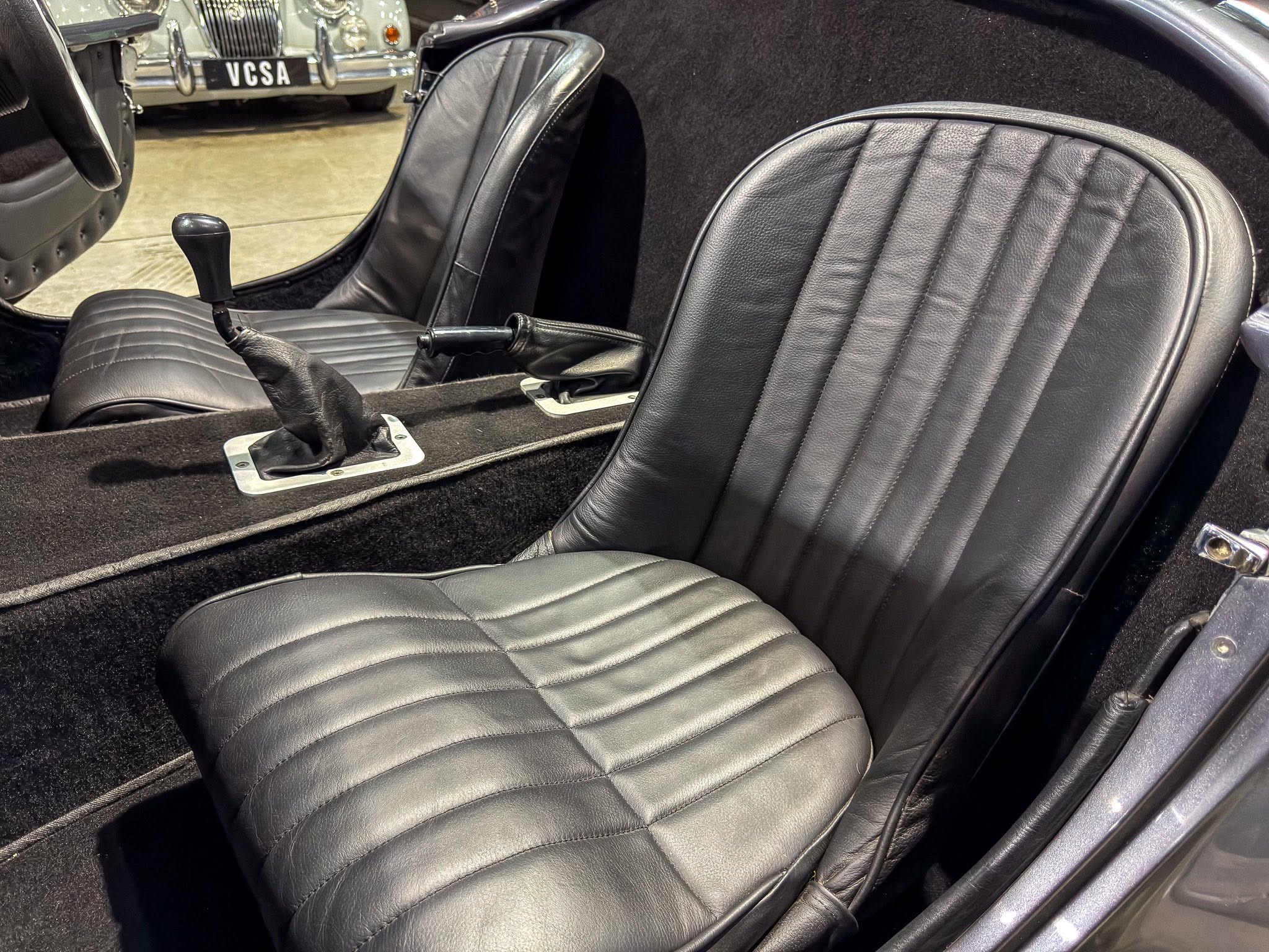 Interior of a vintage car with black ribbed leather seats, a manual gear lever, and a handbrake on a black carpet.