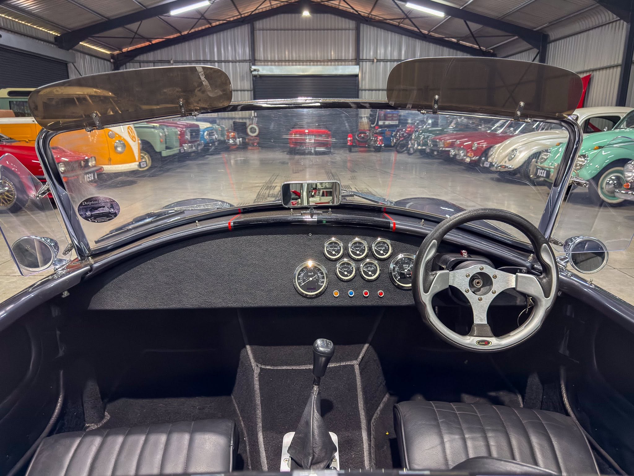View from the driver’s seat of a classic convertible, dashboard gauges visible with a row of vintage cars in a showroom ahead.