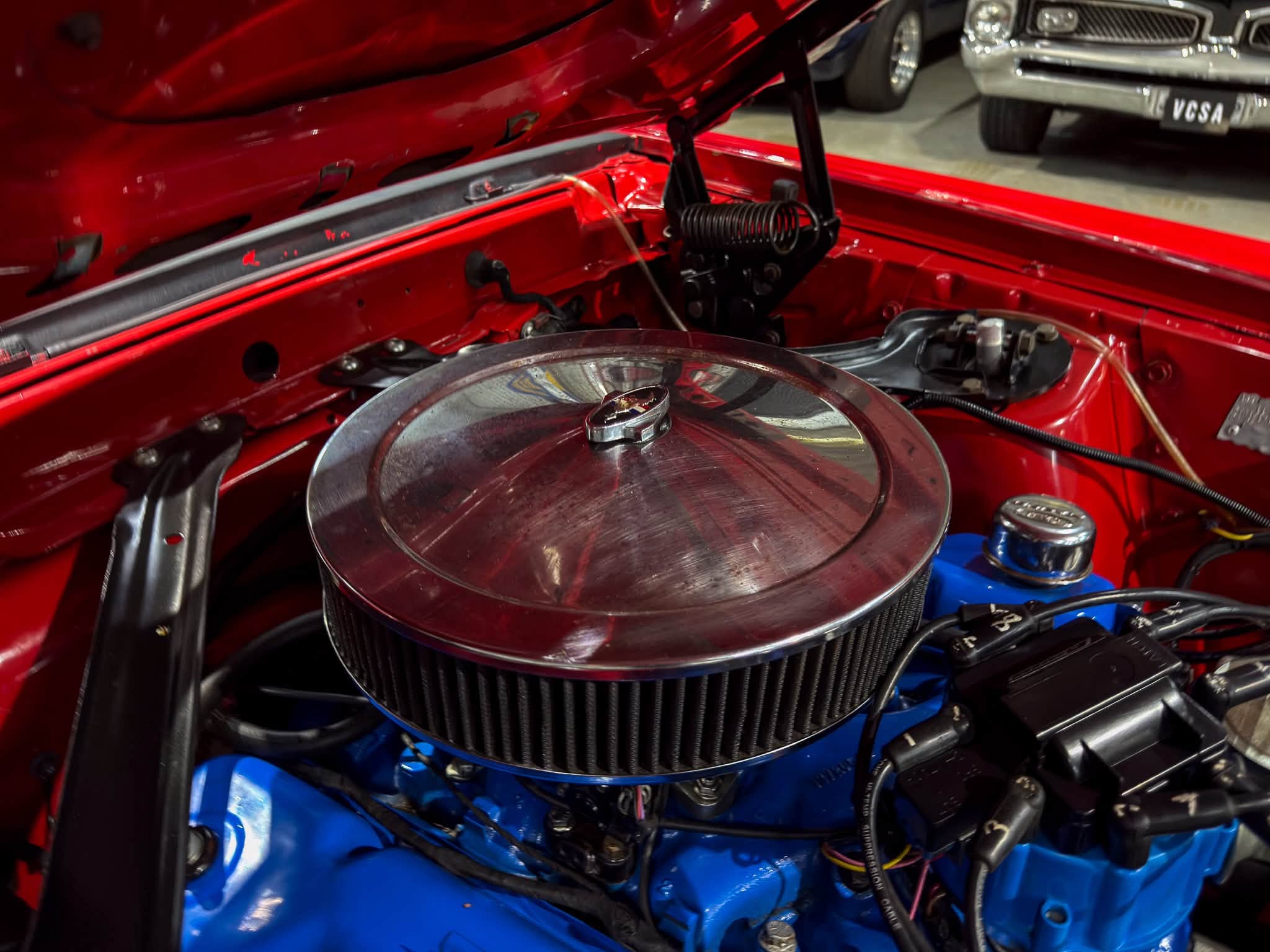 Red car engine bay with a large chrome air filter top centered, blue components below, and exposed hoses and wiring surrounding the firewall.
