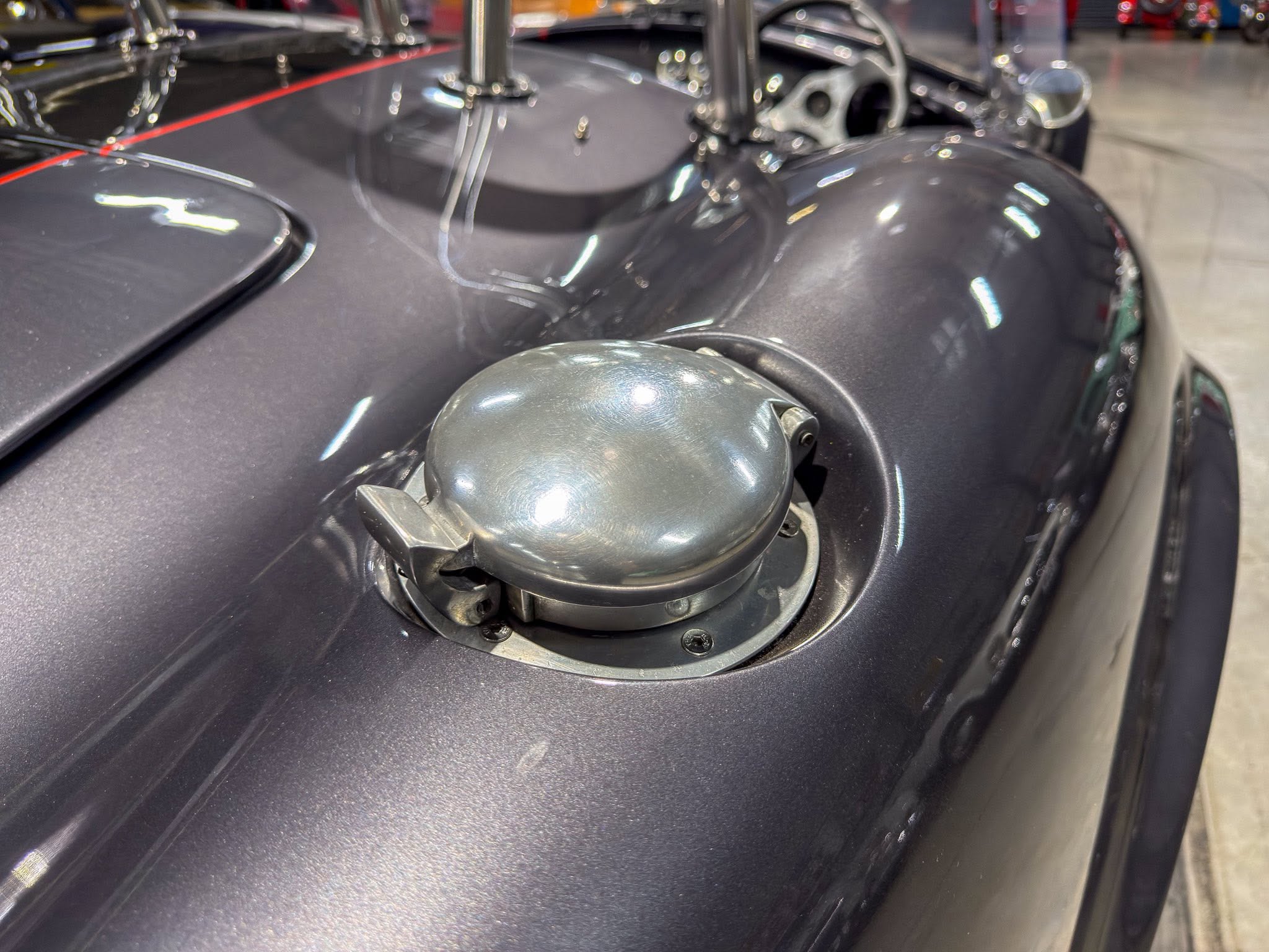 Close-up of a dark, glossy motorcycle fuel tank with a silver/metal gas cap in the foreground; showroom background is blurred.”] ,