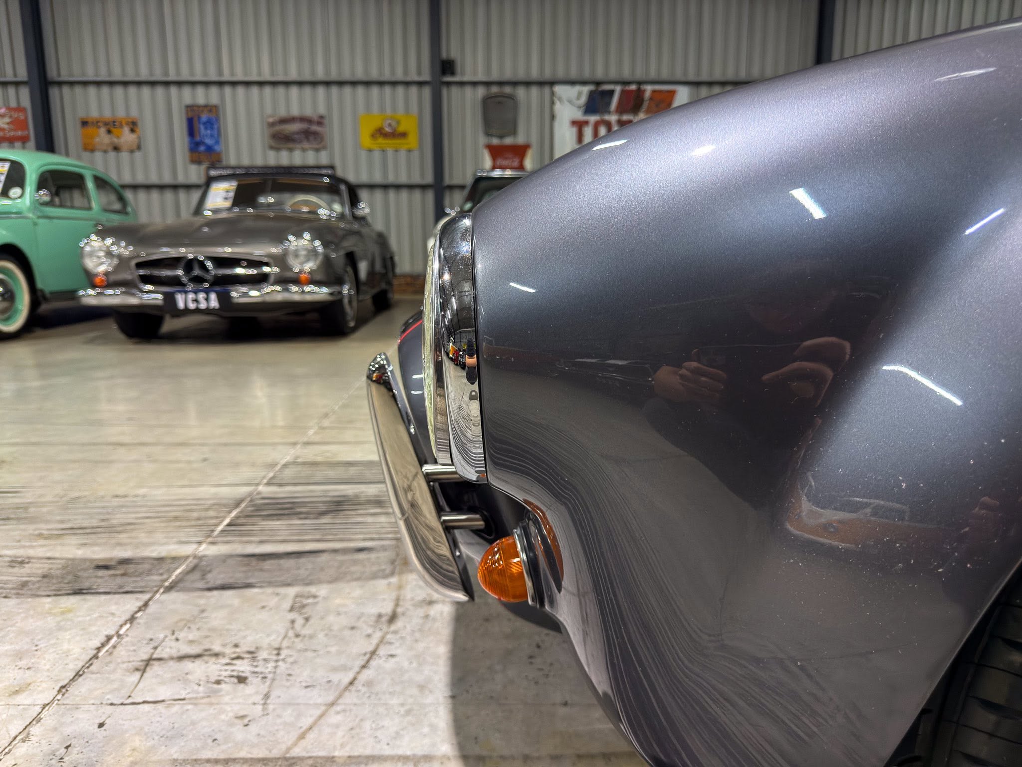 Close-up of a gray vintage car's curved body and orange indicator light, with classic cars in a showroom background.