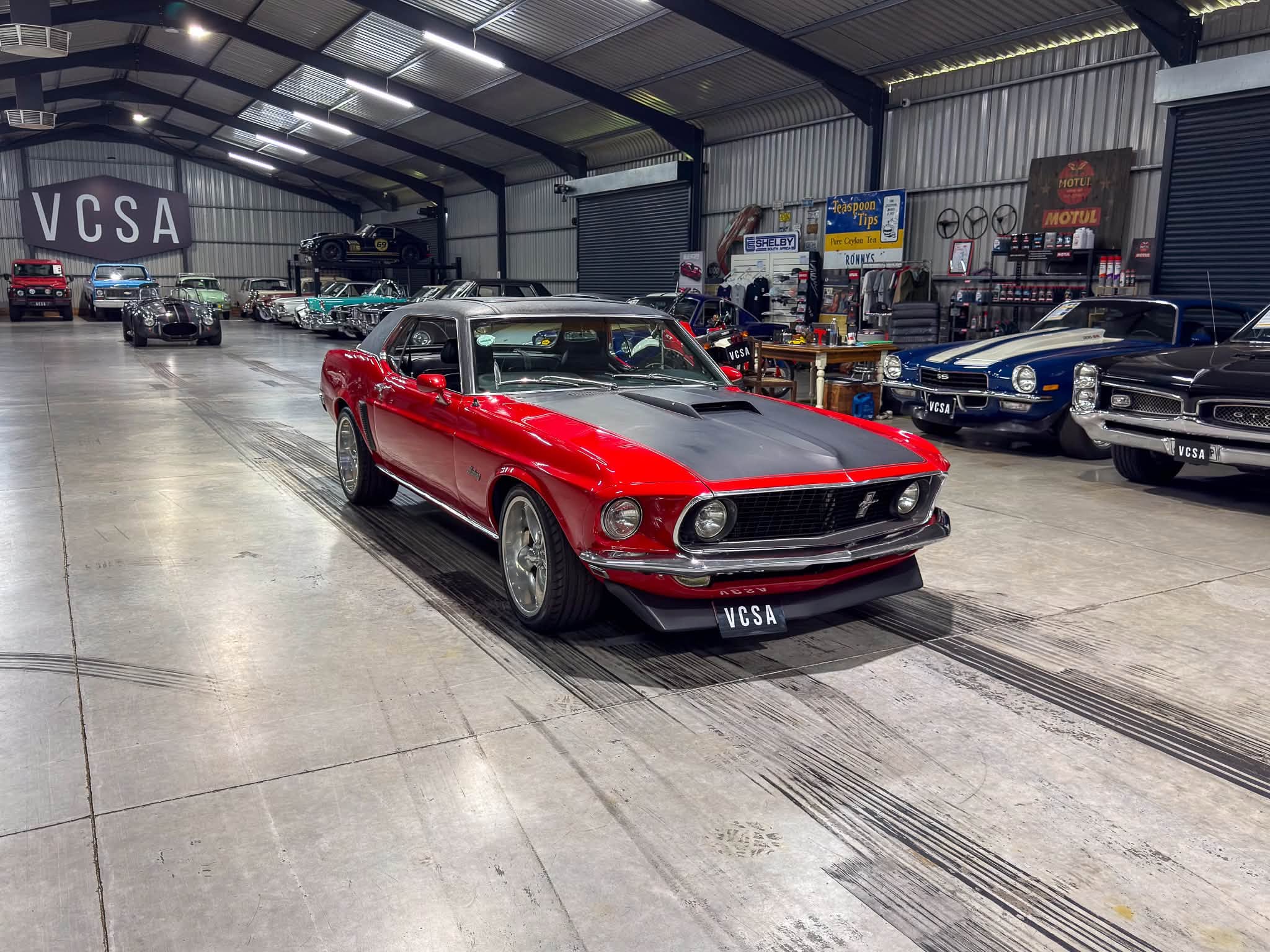 Interior of a classic car showroom with vintage vehicles lined along the walls under industrial lighting, VC SA signage visible in the back.