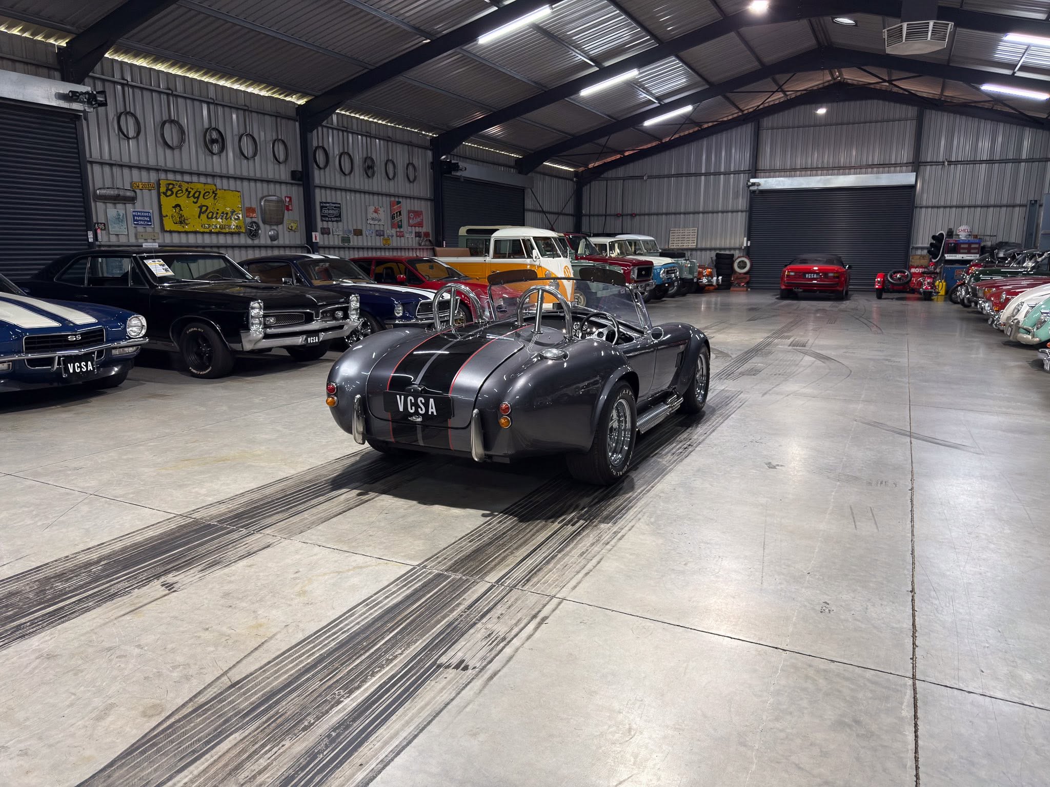 Indoor warehouse showroom filled with vintage cars lined up along the walls, metal siding and ceiling visible.