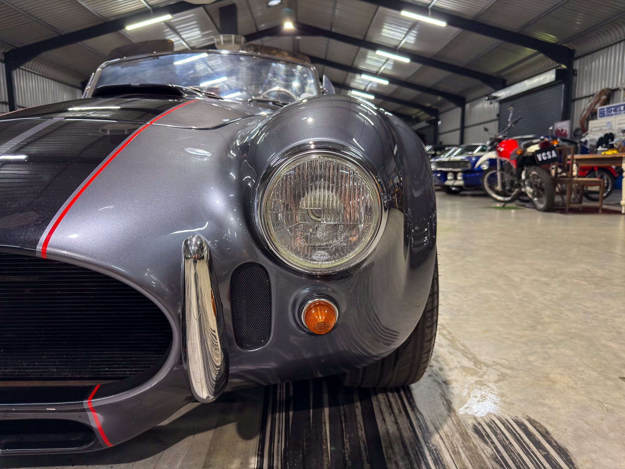 Close-up of a vintage gray sports car front, headlight and red pinstripe, in a workshop with other vehicles in the background.