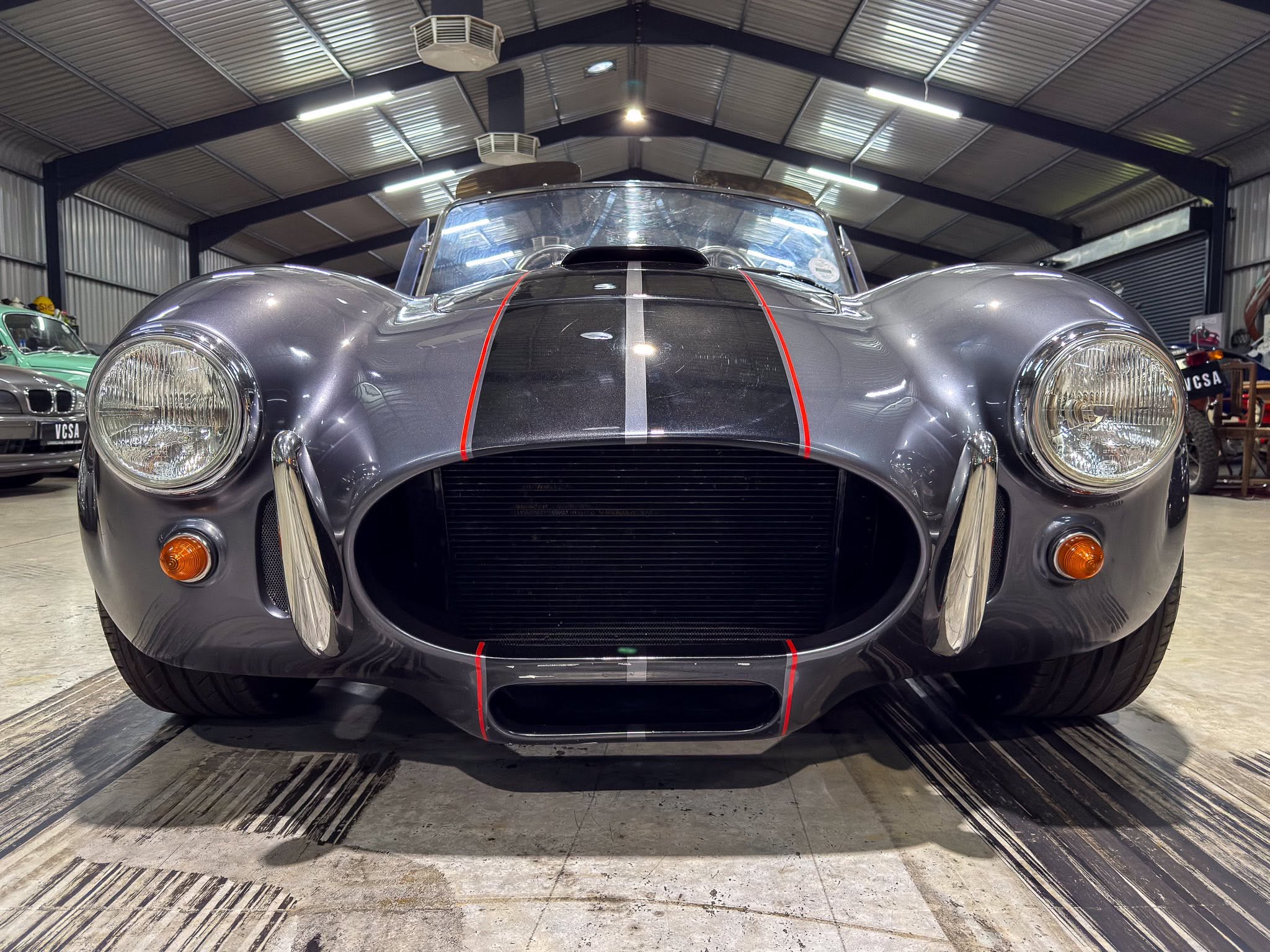 Front view of a vintage silver sports car with red racing stripes, indoors in a metal workshop showroom.